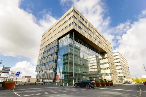 Modern kantoorgebouw met glazen gevel en een geparkeerde auto ervoor onder een bewolkte hemel.