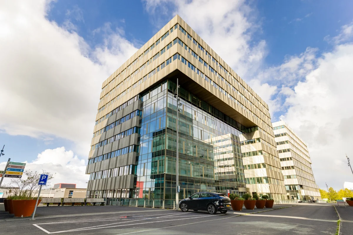 Modern kantoorgebouw met glazen gevel en een geparkeerde auto ervoor onder een bewolkte hemel.