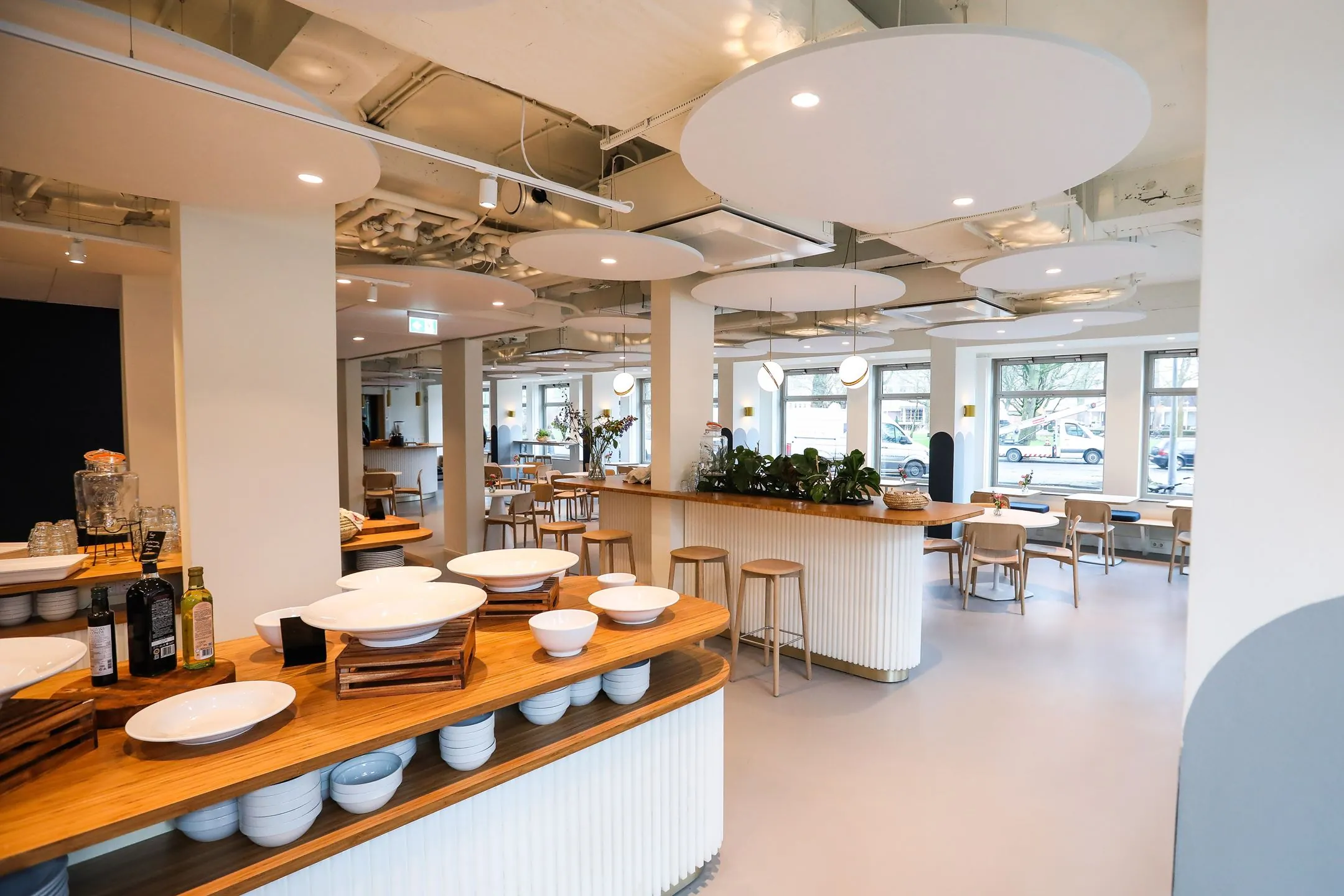 Moderne, open bedrijfskantine met houten meubels, veel lichtinval en een buffetopstelling met witte schalen.