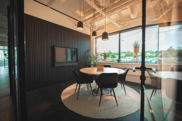 Moderne vergaderruimte met ronde tafel, vijf stoelen, een tv-scherm en grote ramen met uitzicht op een parkeerterrein.