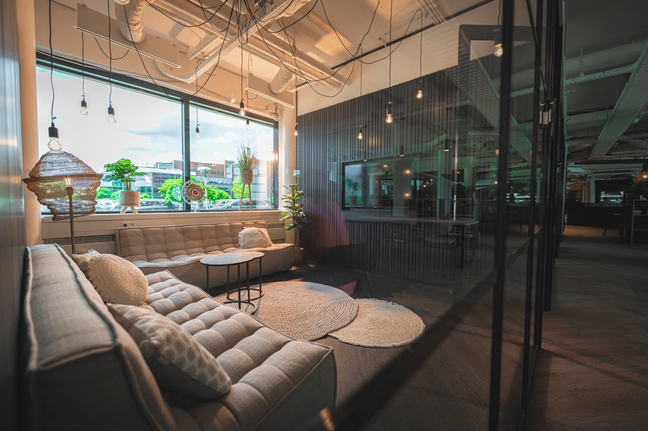 Moderne loungehoek in een kantoorruimte met comfortabele banken, hanglampen en grote ramen met uitzicht op geparkeerde auto's.