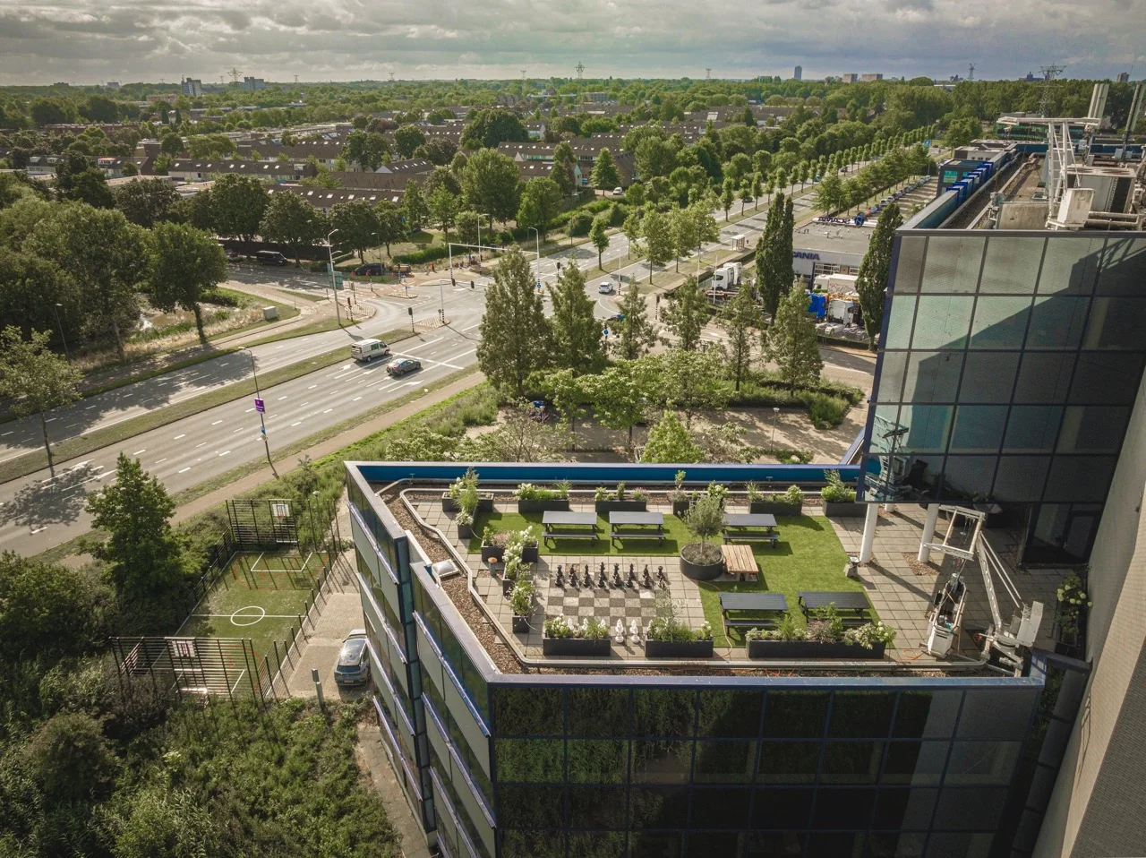 Dakterras met zitgelegenheid en buitenschaakspel op een modern kantoorgebouw aan een drukke weg, omringd door veel groen.
