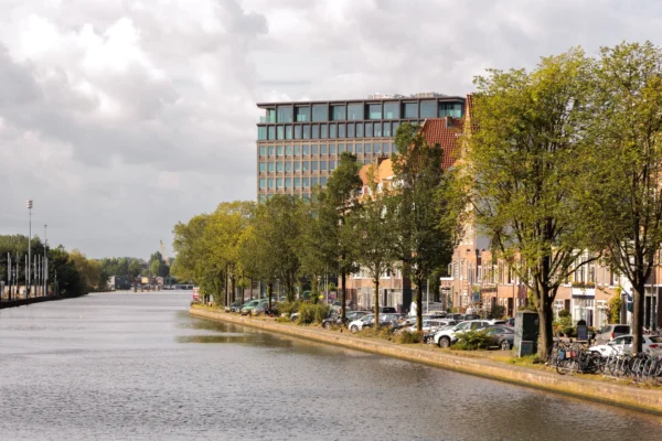 Uitzicht op een schilderachtige kade met bomen, geparkeerde auto's en een modern gebouw op de achtergrond.