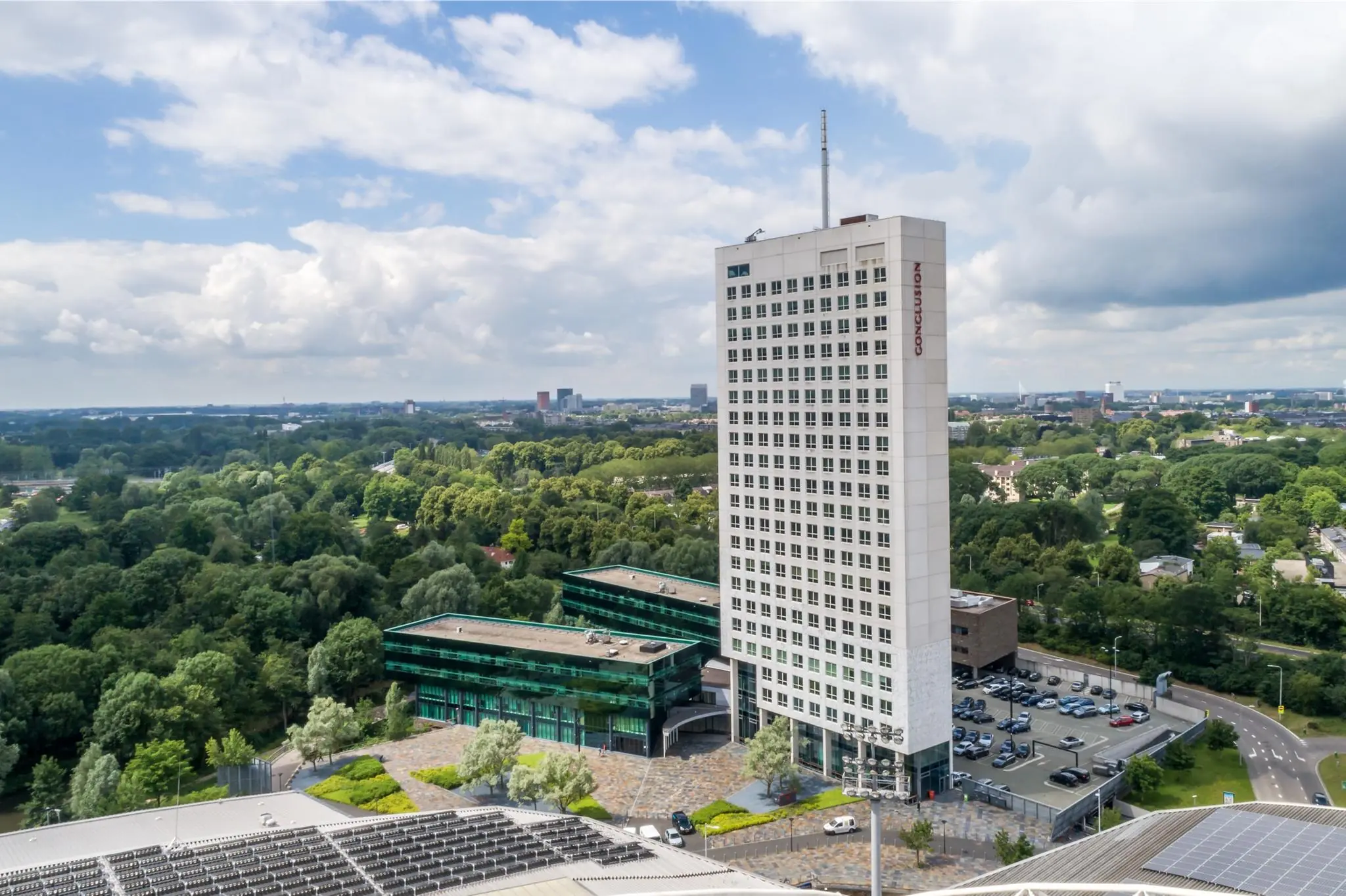 Luchtfoto van de kantoortoren ‘Conclusion’ aan het Herculesplein in Utrecht, omringd door bomen en parkeerterreinen.