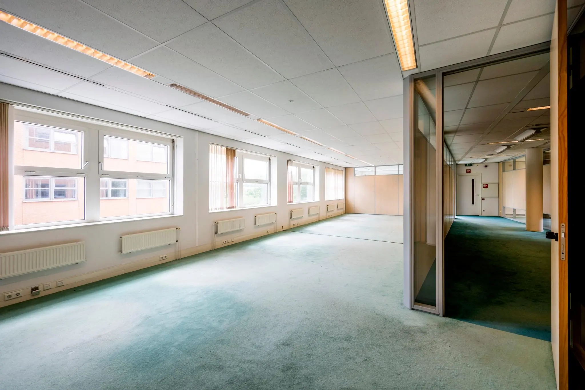 Licht en ruim kantoor met grote ramen, systeemplafond en groen tapijt.
