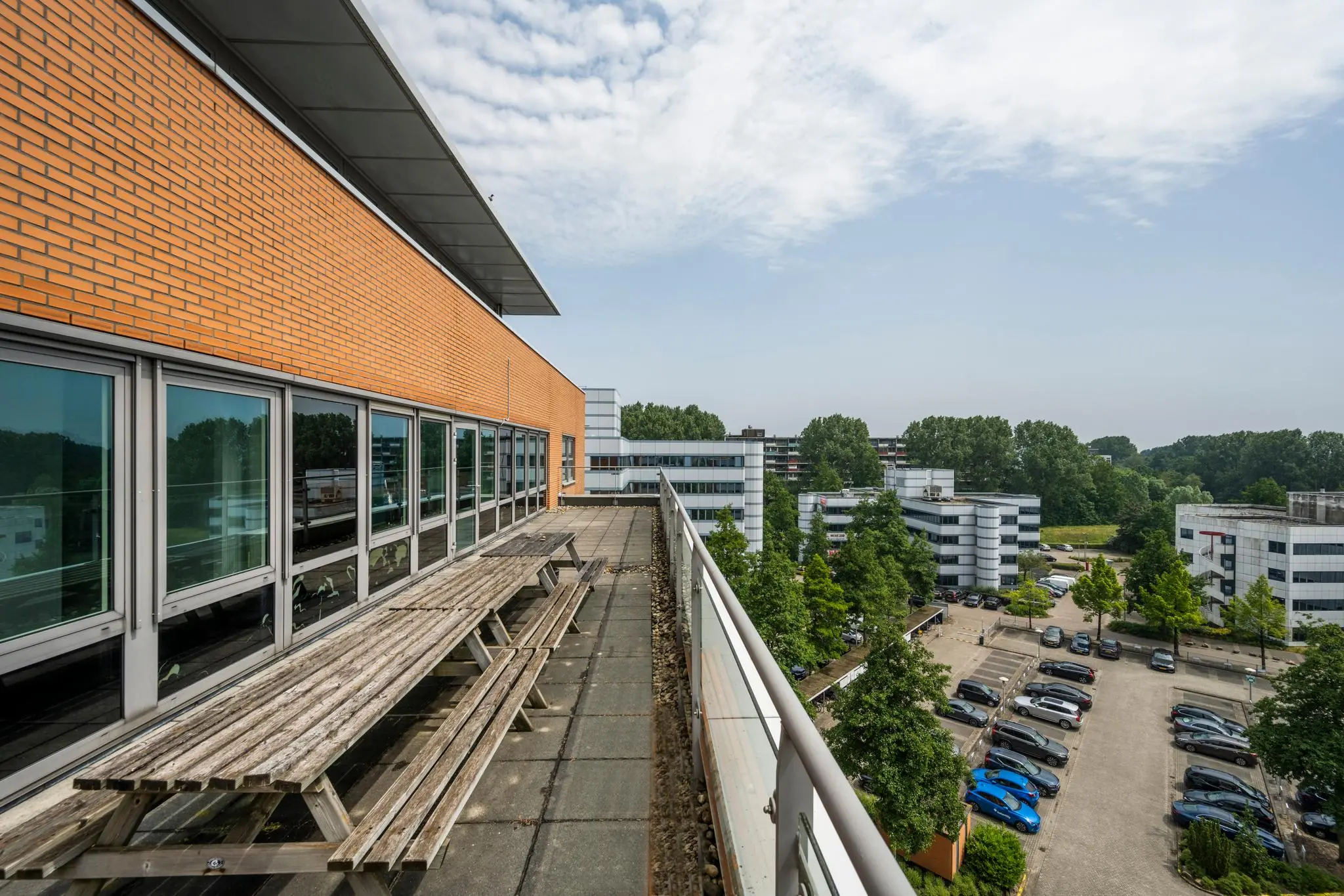 Buitenruimte met picknicktafels op een balkon van een modern kantoorpand, uitkijkend over een parkeerterrein en omliggende kantoorgebouwen.