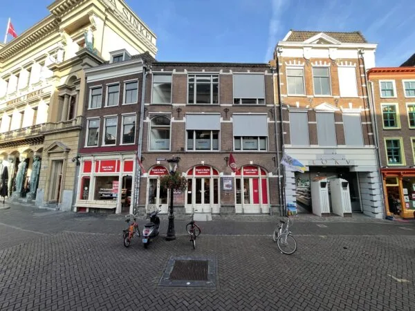 Een straatbeeld van de Ganzenmarkt met historische gebouwen en geparkeerde fietsen op een zonnige dag.