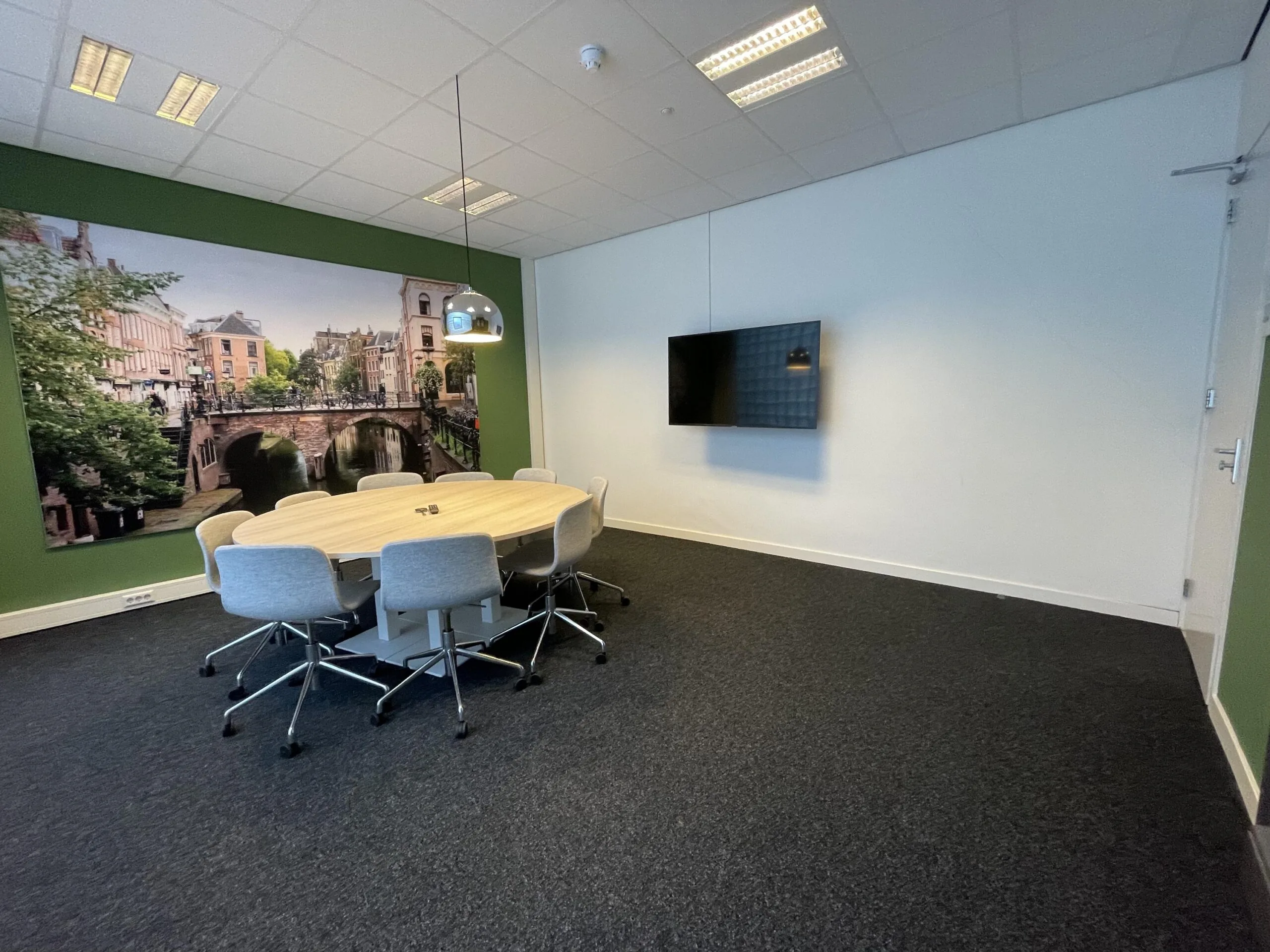 Een vergaderruimte met een ovale tafel, stoelen, een muur met stadsafbeelding en een scherm aan de wand.