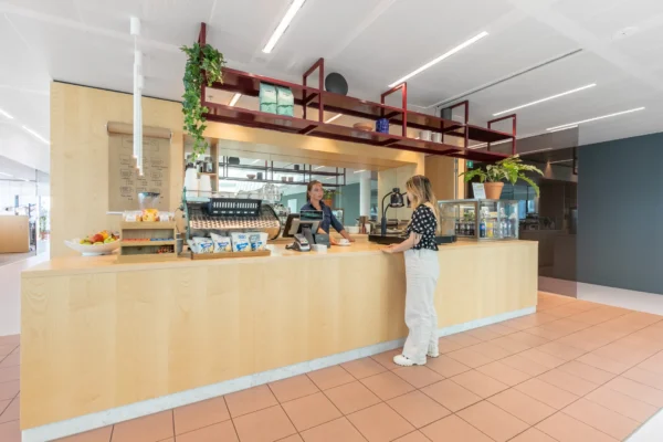 Een vrouw staat aan de balie van een modern café en spreekt met een medewerkster.