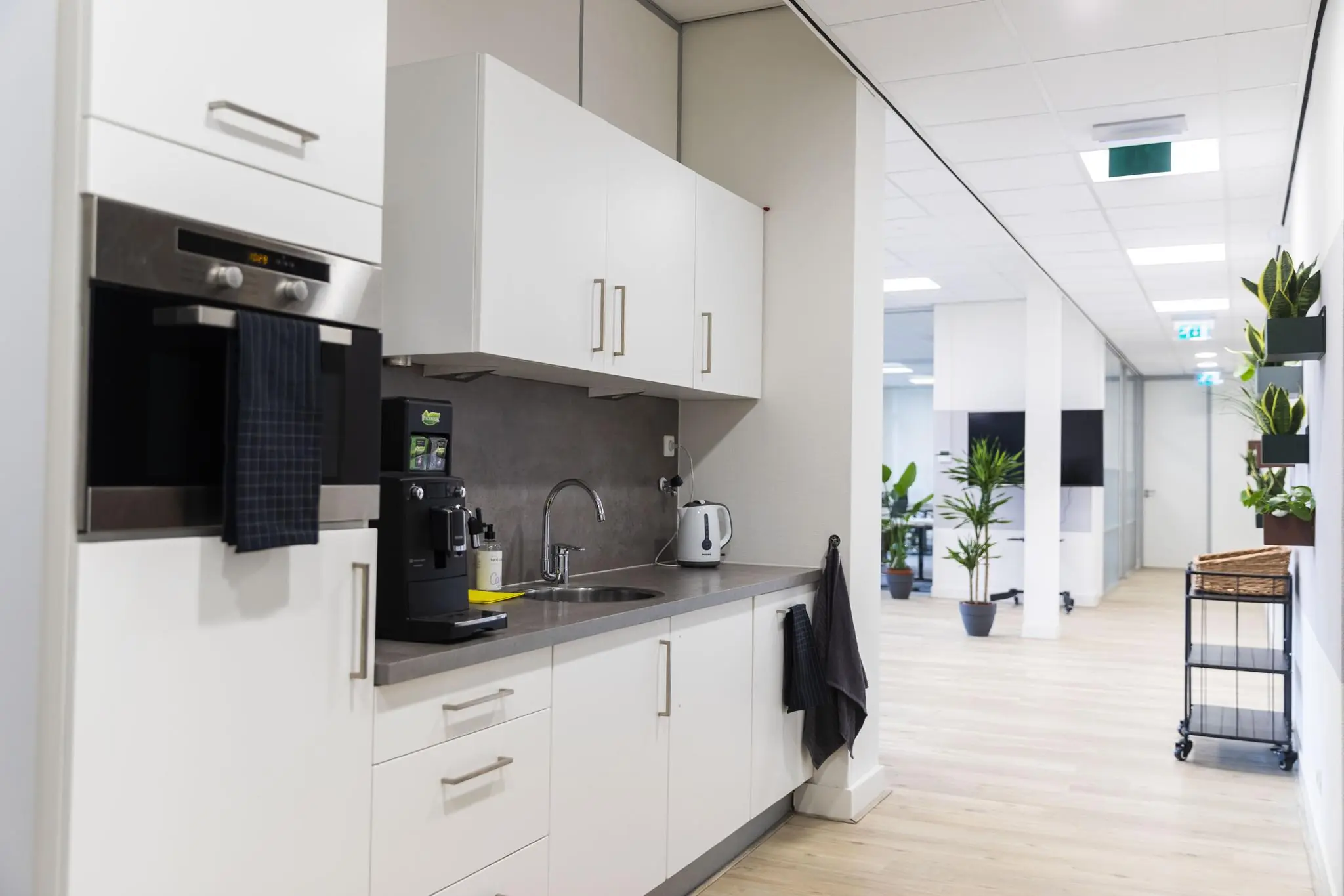 Moderne kantoorpantry met witte kasten, koffiezetapparaat en uitzicht op een lichte gang met planten.