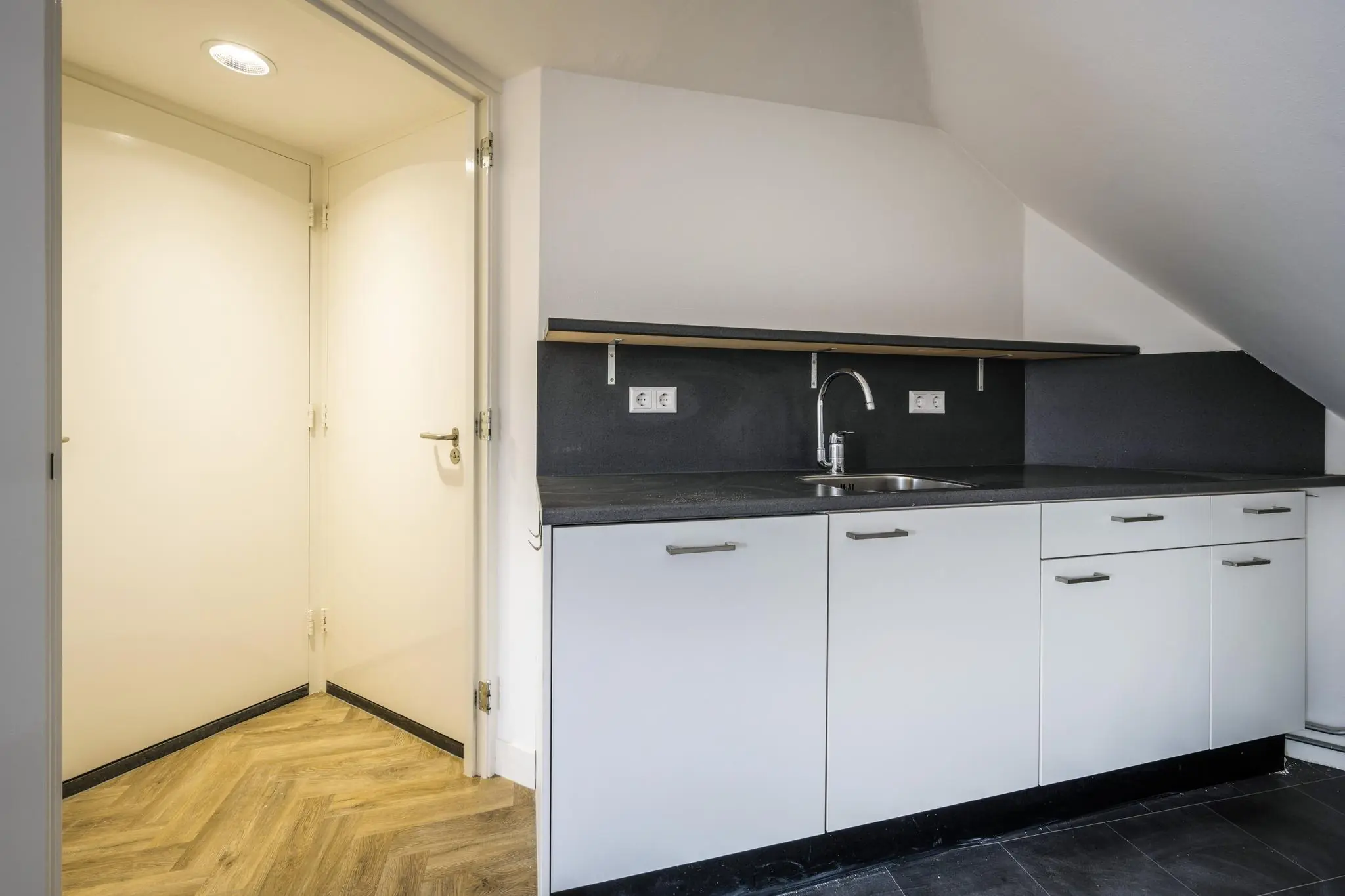 Moderne kitchenette met witte kasten, zwarte blad en aangrenzende hal met houten visgraatvloer.