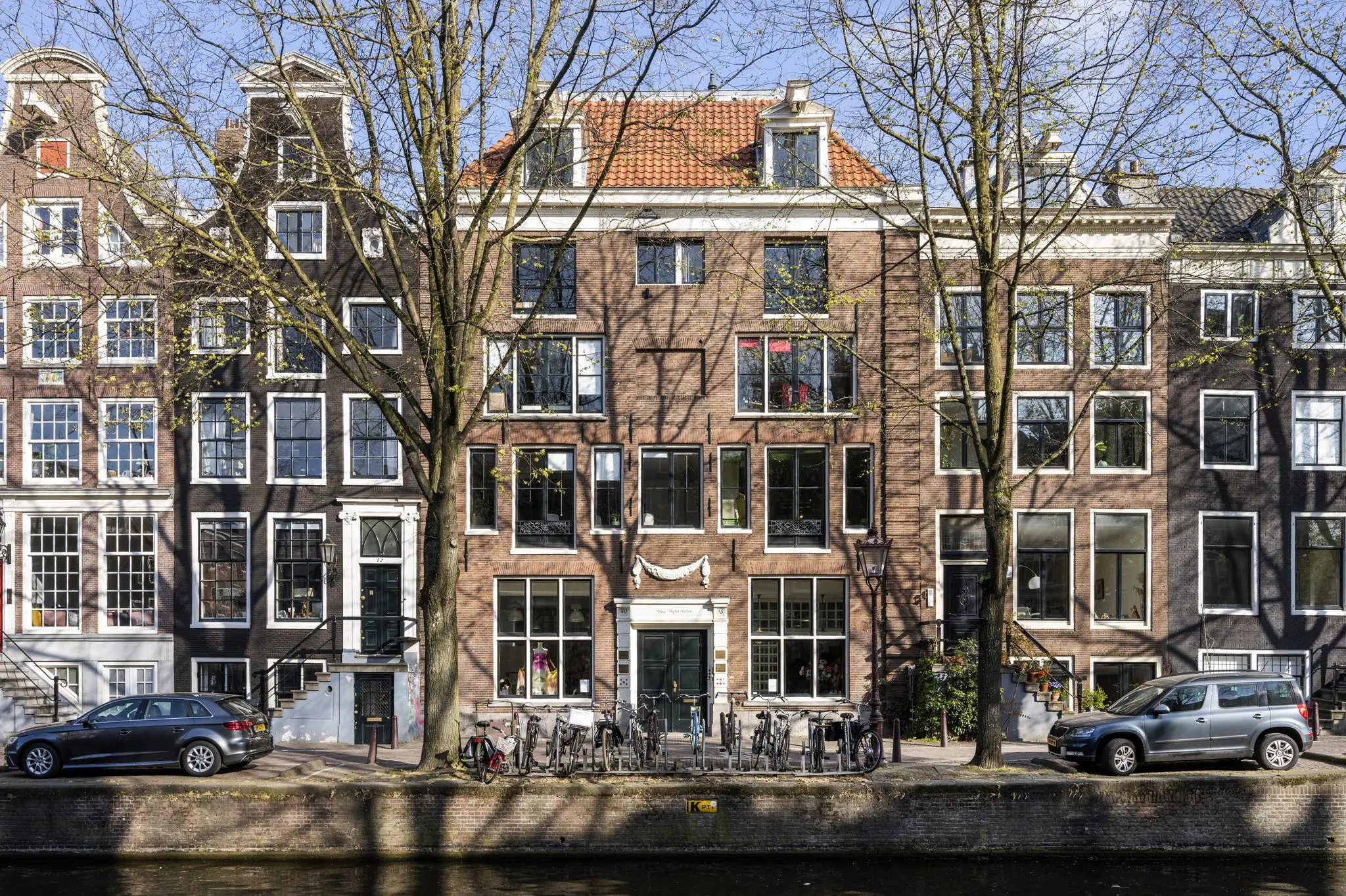 Gevels van historische grachtenpanden aan de Leidsegracht in Amsterdam met bomen, fietsen en geparkeerde auto's langs het water.