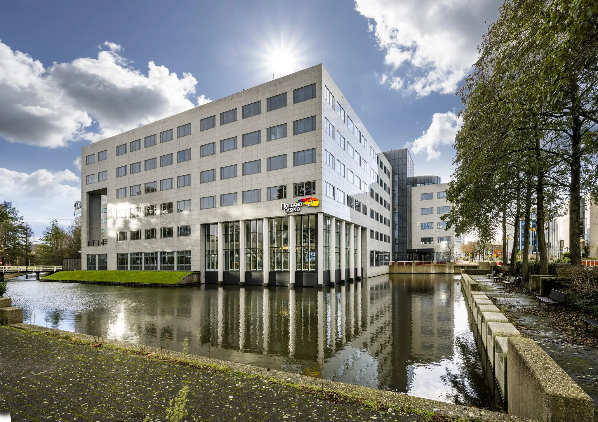 Modern kantoorgebouw aan de Neptunusstraat met Holland Casino-logo, gelegen aan een waterpartij onder een deels bewolkte lucht.