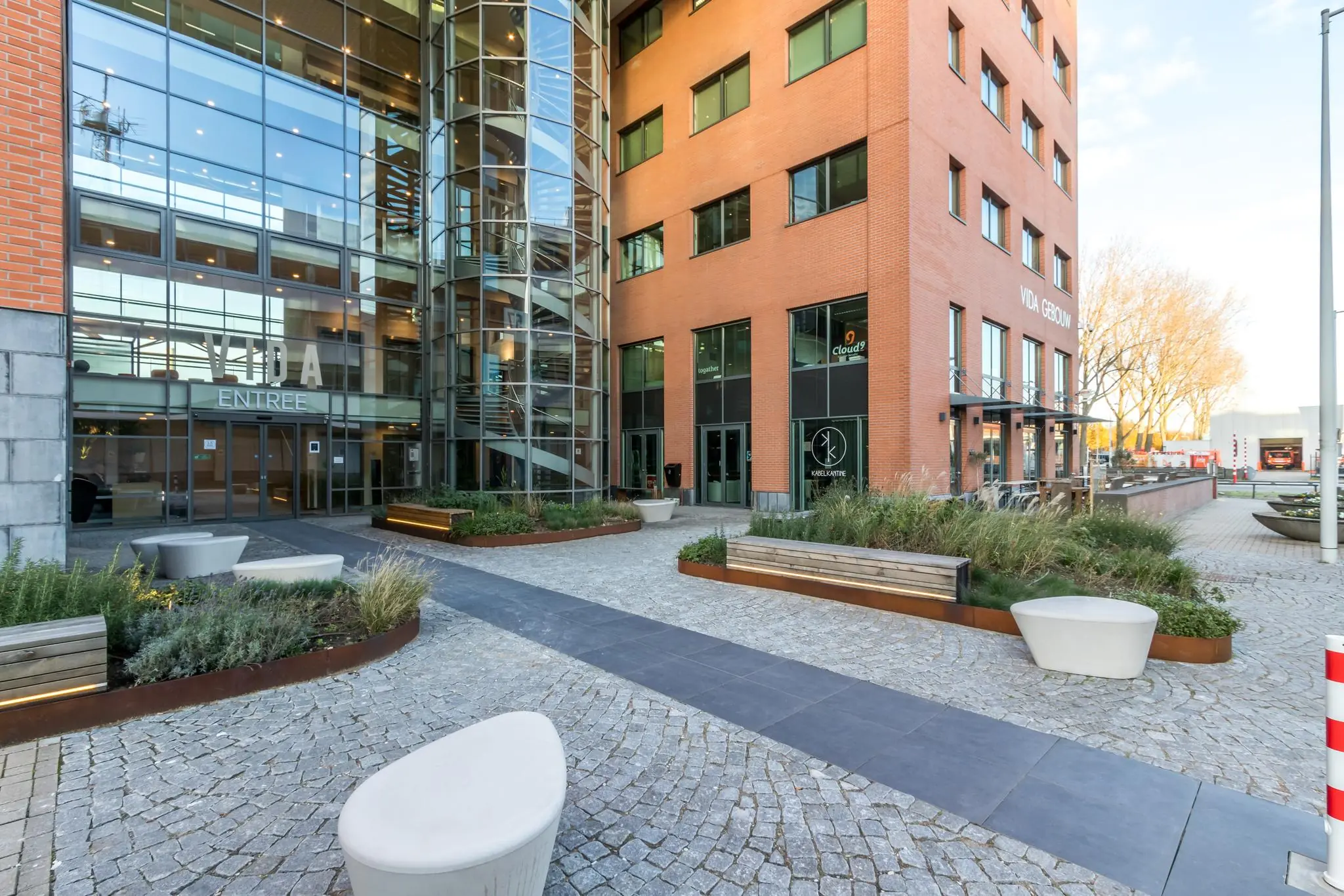 Ingang van het VIDA-gebouw aan de Kabelweg met moderne gevel, glazen entree en aangelegde buitenruimte met zitplekken.