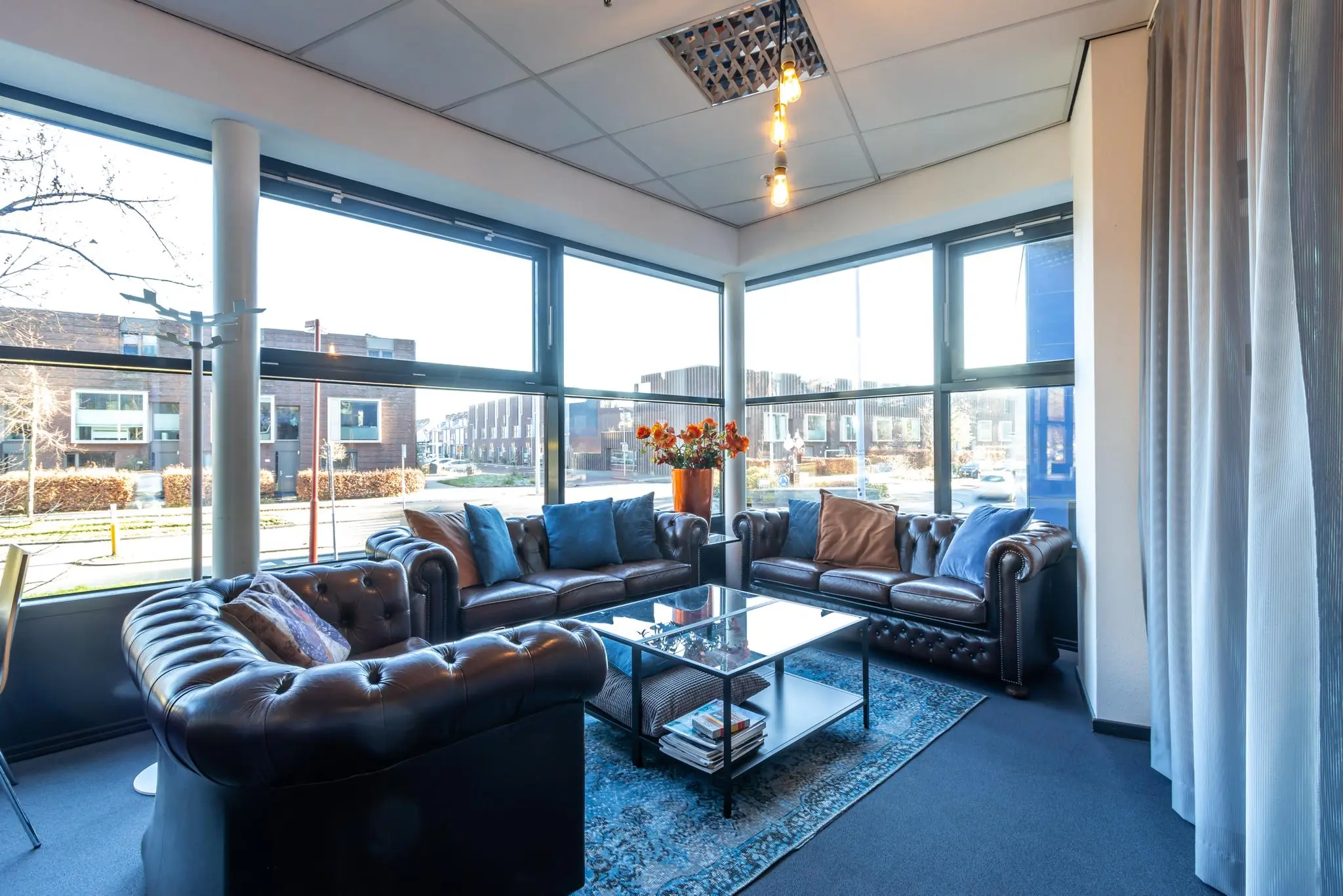 Zonnige wachtruimte met leren Chesterfield-banken, glazen salontafel en uitzicht op de straat aan de Oudlaan.