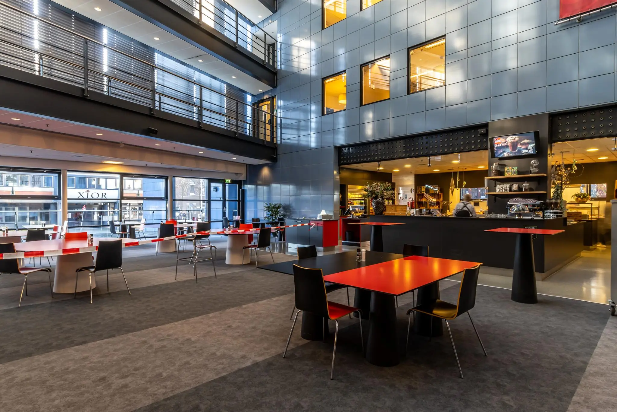 Moderne, open ontmoetingsruimte met rood-zwarte tafels, hoge plafonds en een koffiebar in gebouw aan de Oudlaan.