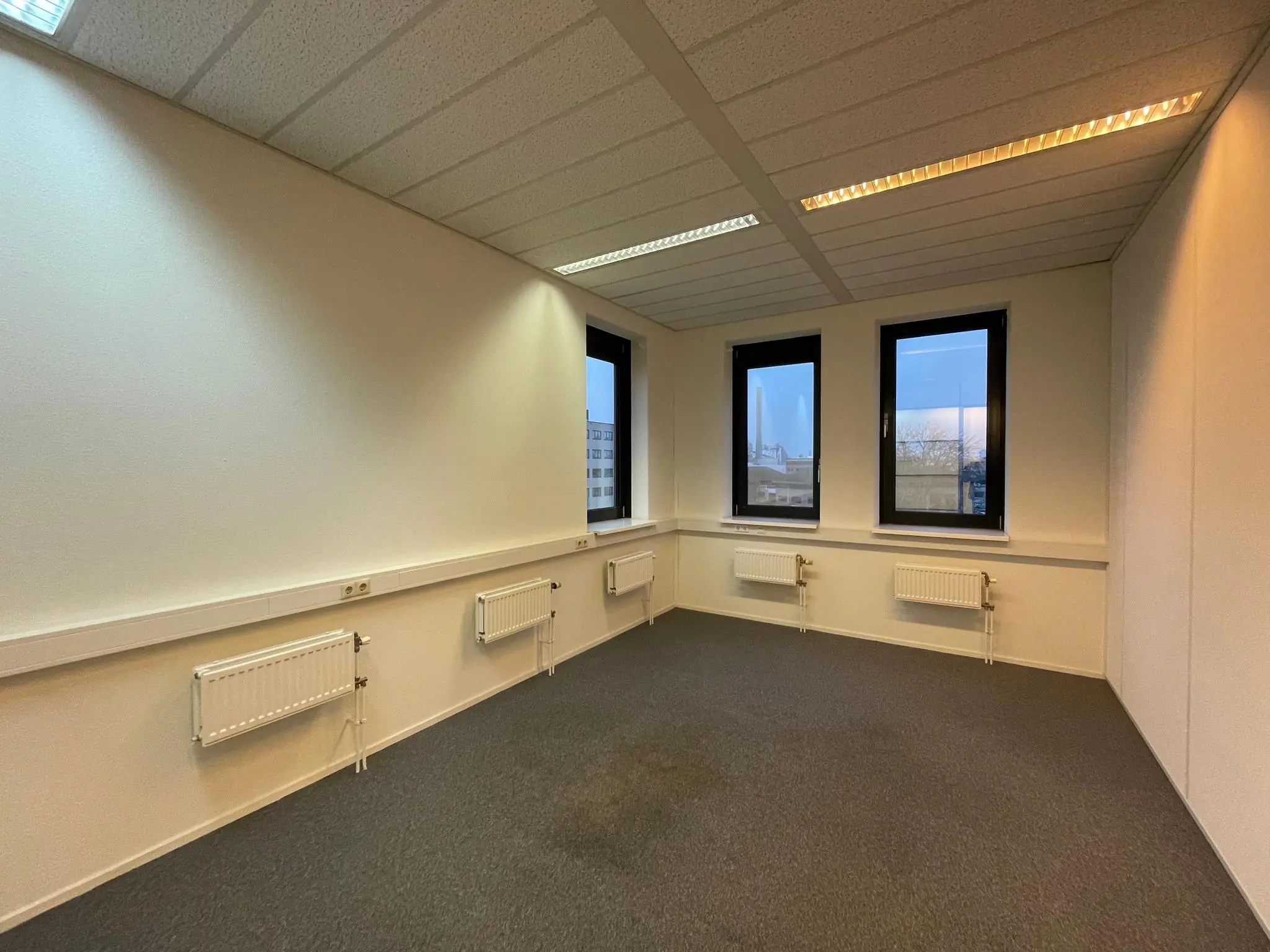 Leeg kantoorkantoor aan de Zekeringstraat met drie ramen, grijze vloerbedekking en witte muren met radiatoren.