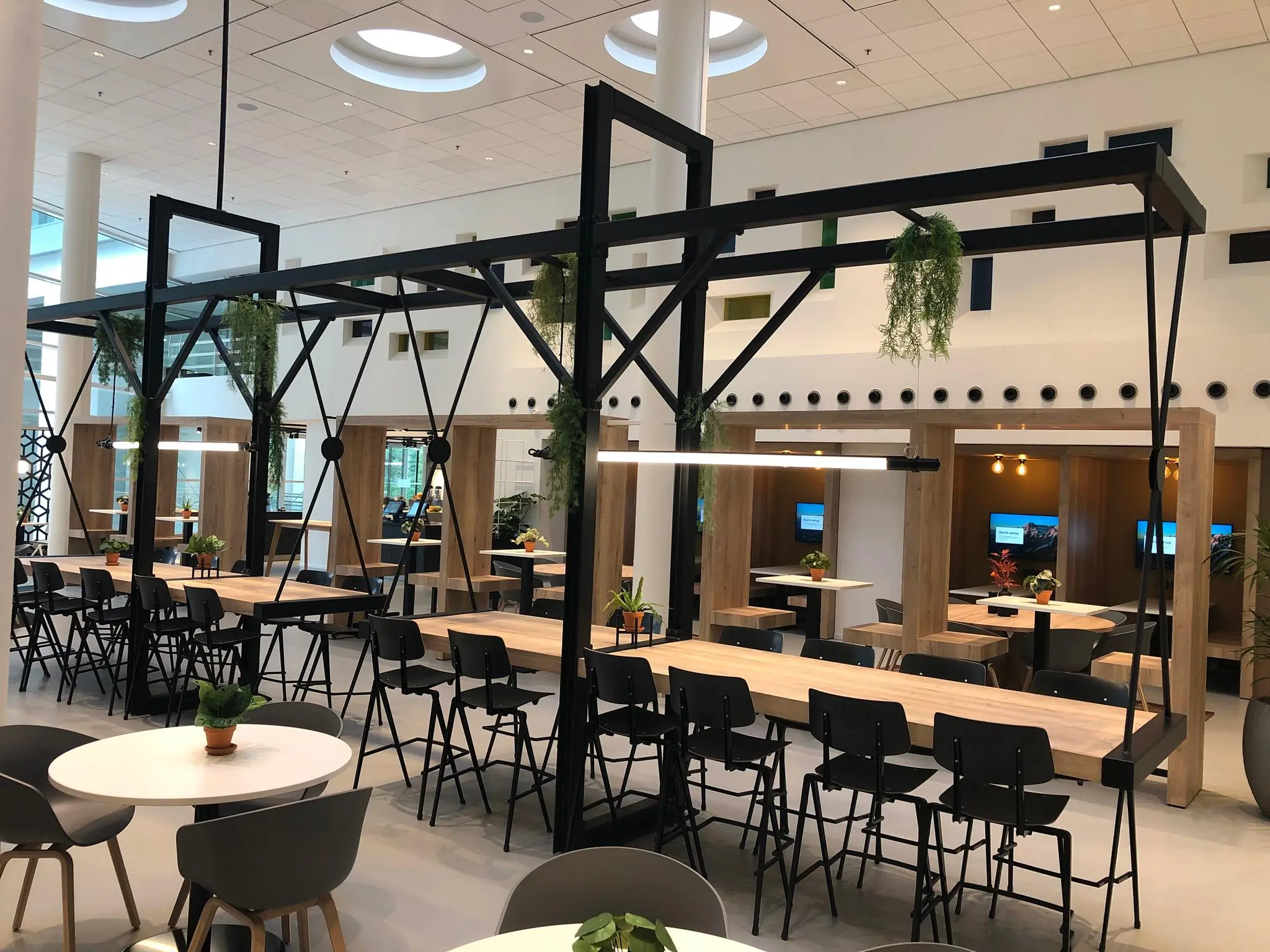 Moderne en open kantoorkantine aan de Newtonlaan met lange houten tafels, zwarte stoelen, hangplanten en werkplekken met schermen.