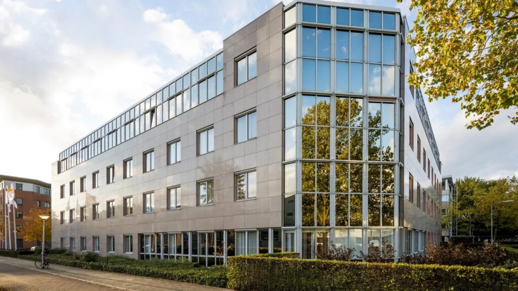 Modern kantoorgebouw met glazen gevel en reflectie van bomen in de ramen, genaamd Wisselwerking.