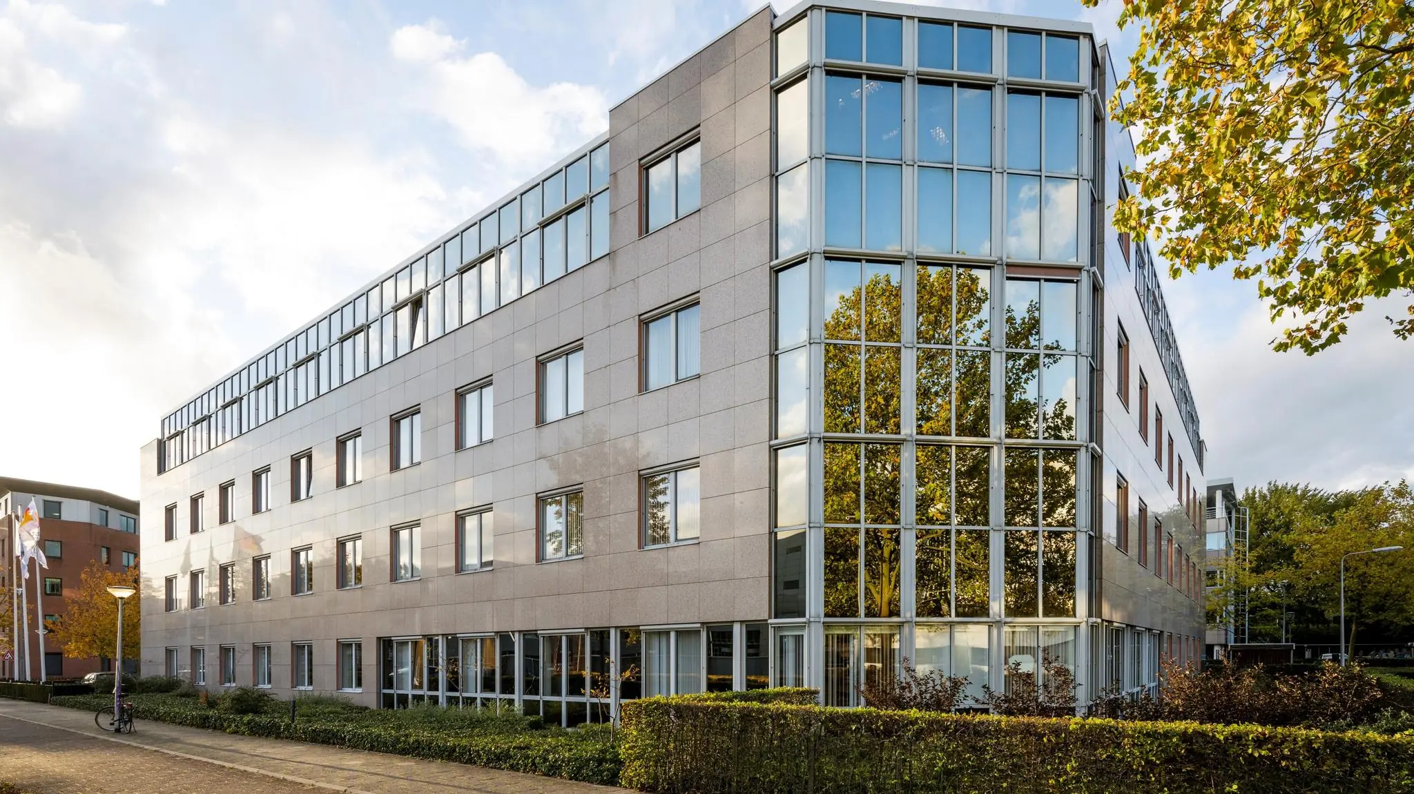 Modern kantoorgebouw met glazen gevel en reflectie van bomen in de ramen, genaamd Wisselwerking.