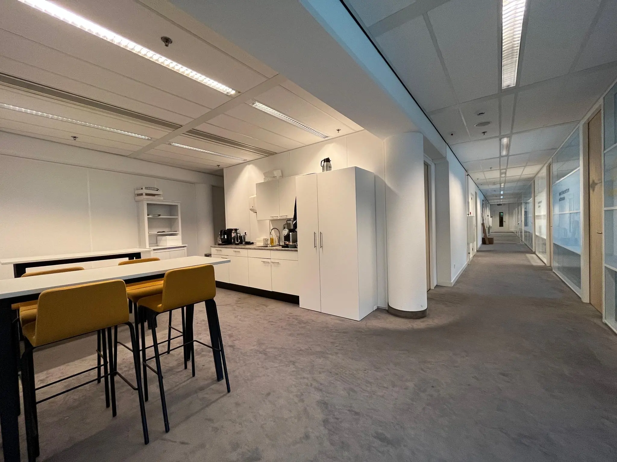 Kantoorruimte op Herculesplein met een moderne pantry en gele barkrukken naast een gang met glazen kantoorwanden.