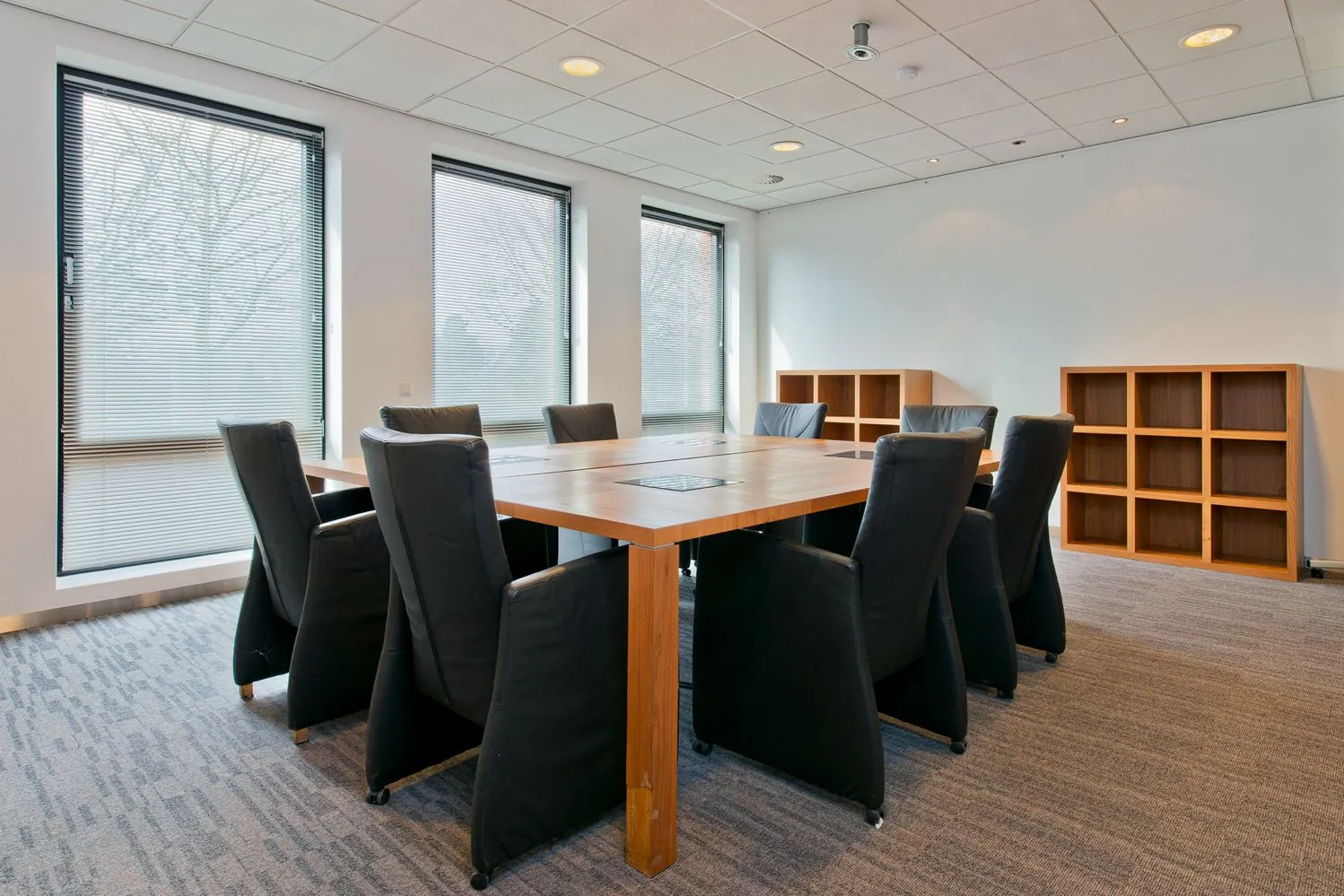 Een vergaderruimte met een grote houten tafel, zwarte stoelen en kasten, met zicht op drie ramen met jaloezieën.