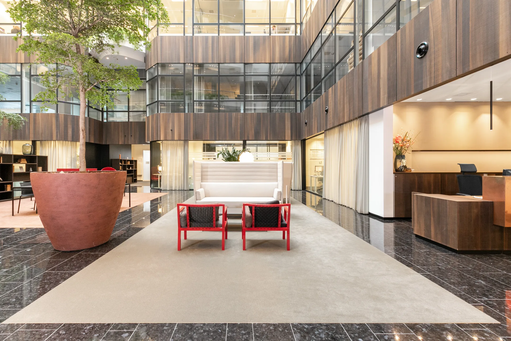 Een moderne lobby met een grote plantenbak, witte bank en rode stoelen op een tapijt, omgeven door glazen wanden en houten panelen.