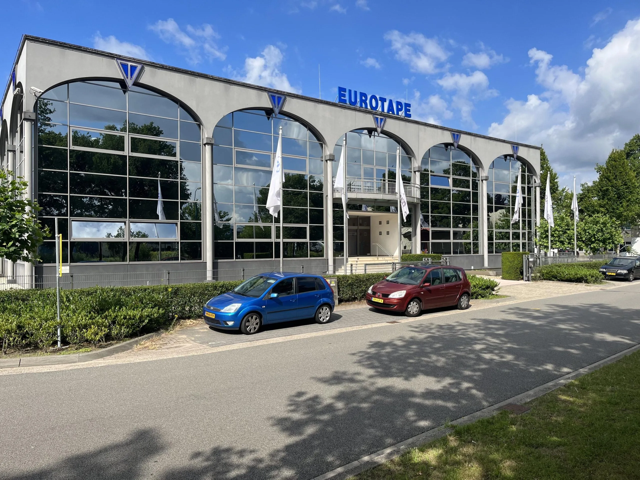 Voorzijde van een modern kantoorpand met de naam "EUROTAPE" erop en geparkeerde auto's op de oprit.