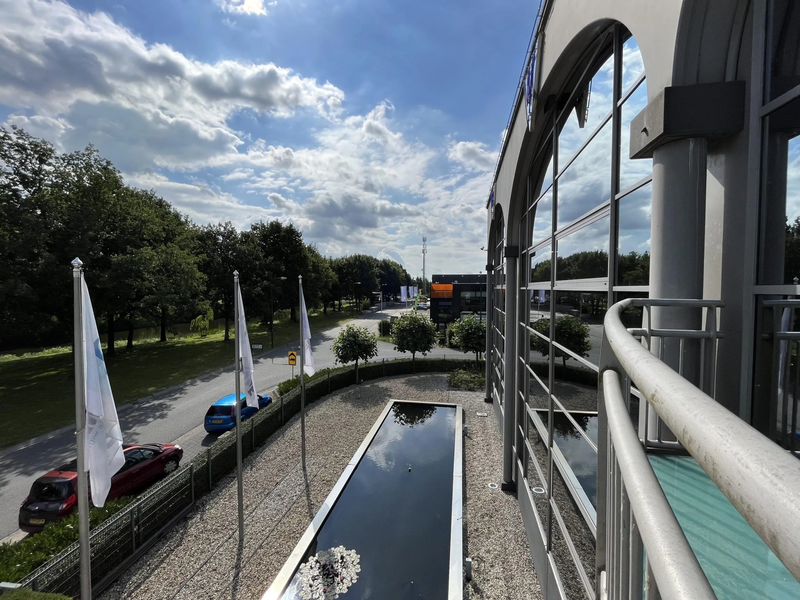 Uitzicht vanaf een gebouw langs de Oostergracht met een vijver, vlaggen en een weg met auto's onder een bewolkte hemel.