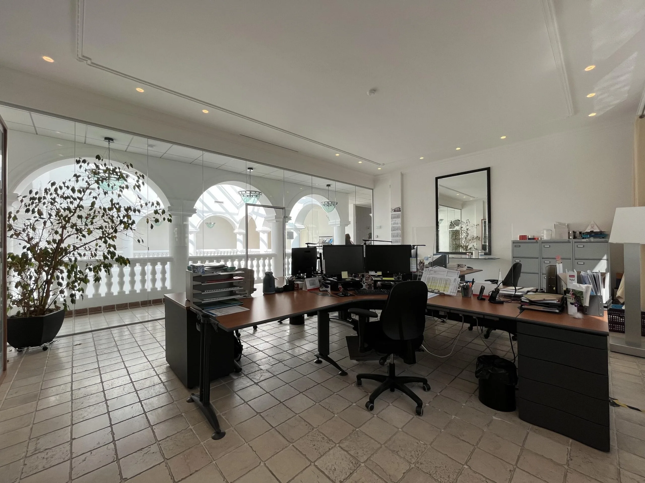 Een modern kantoor met bureaus, stoelen, en computers, met glazen wanden die uitkijken op een marmeren balkon met bogen.