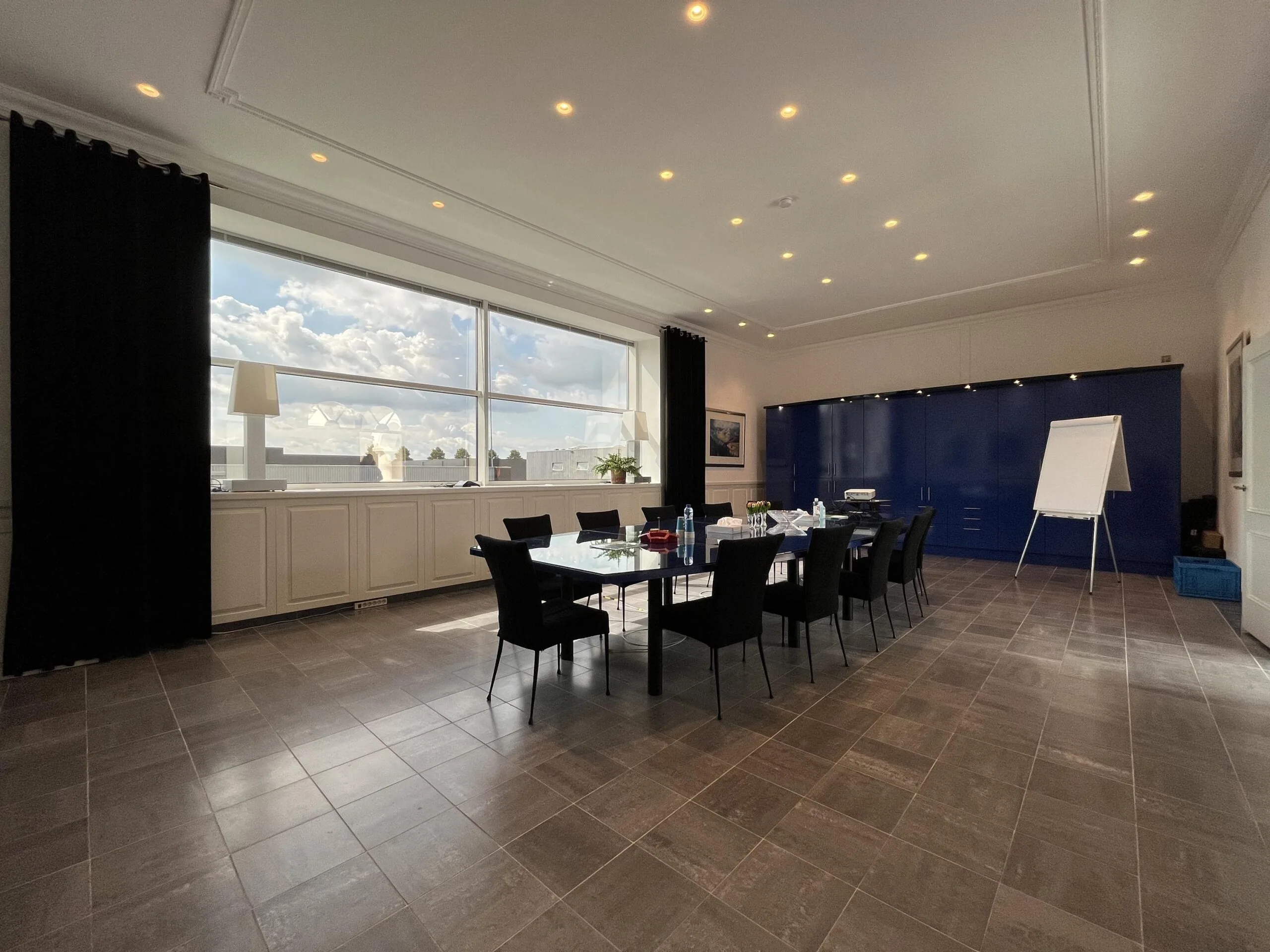 Vergaderruimte met een grote tafel, stoelen, flip-over, en een raam met uitzicht op een heldere lucht.