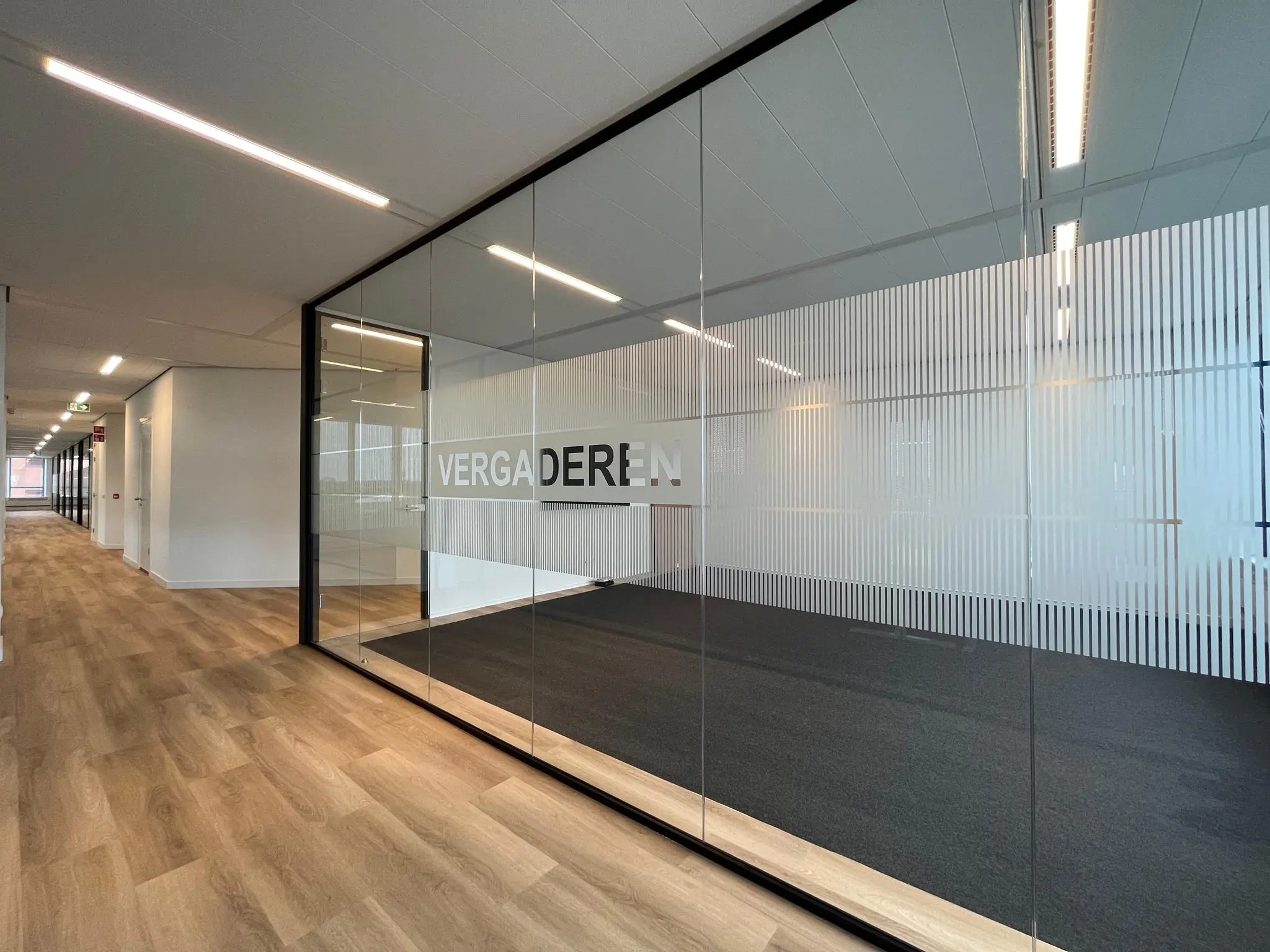 Glazen vergaderruimte met het woord "VERGADEREN" op de wand in een modern kantoorpand aan het Kosterijland.