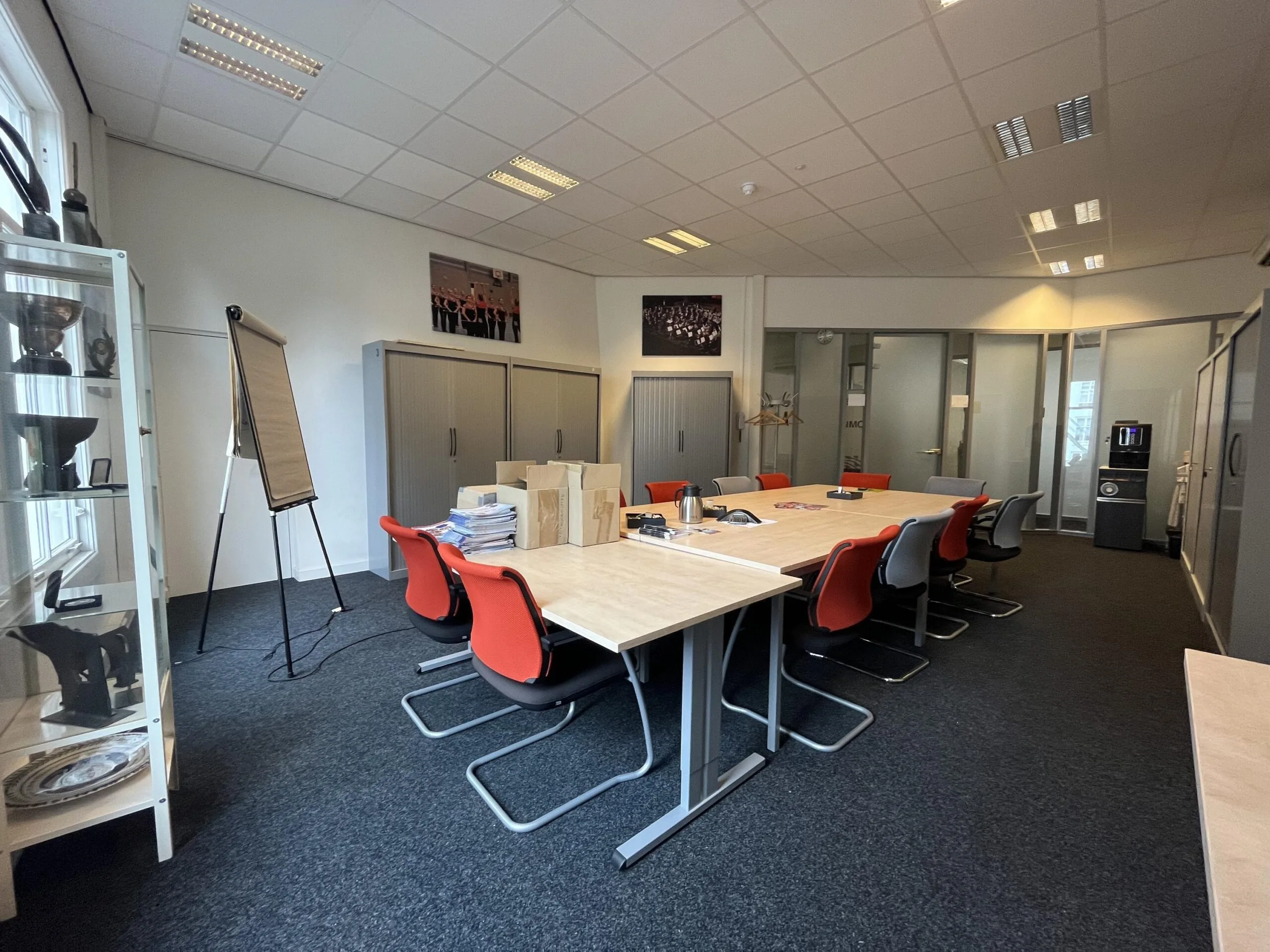 Een vergaderruimte met een lange tafel, rode en grijze stoelen, kastenaankleding en een flip-over.