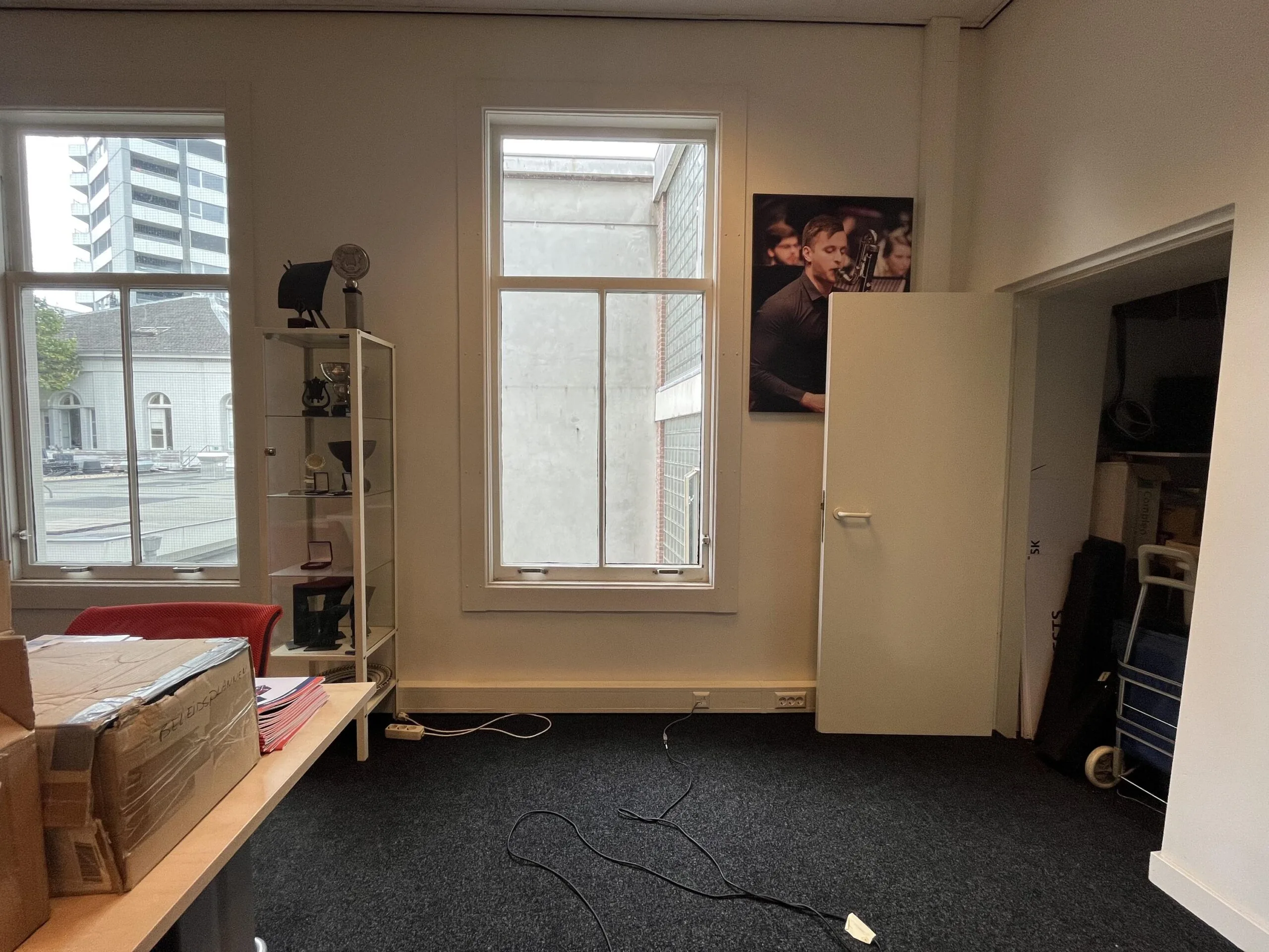 Een kantoorruimte met een bureau, glazen kast, en een openstaande deur naar een opslagruimte, met buitenzicht op een gebouw via twee ramen.