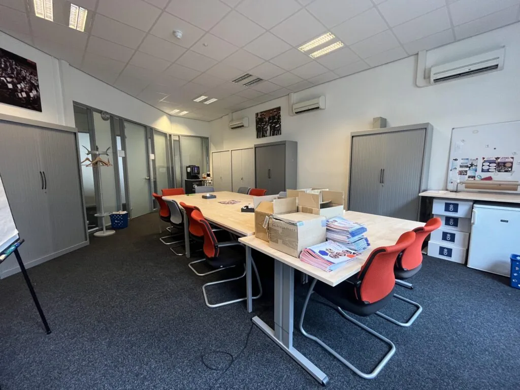 Een vergaderruimte met een grote tafel omringd door rode stoelen, met stapels folders erop en kasten langs de muren.