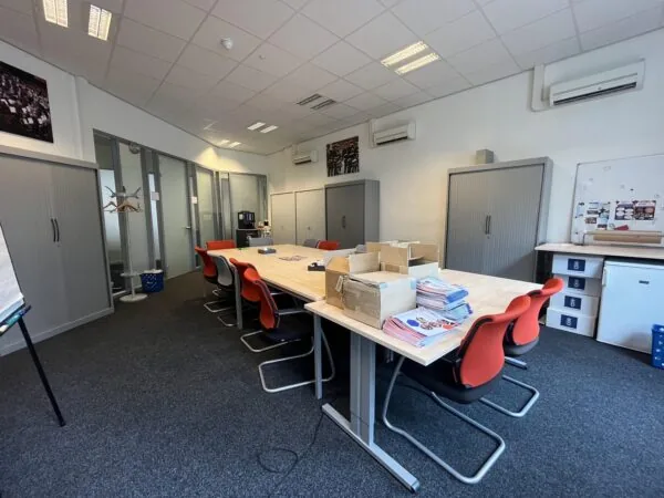 Een vergaderruimte met een grote tafel omringd door rode stoelen, met stapels folders erop en kasten langs de muren.