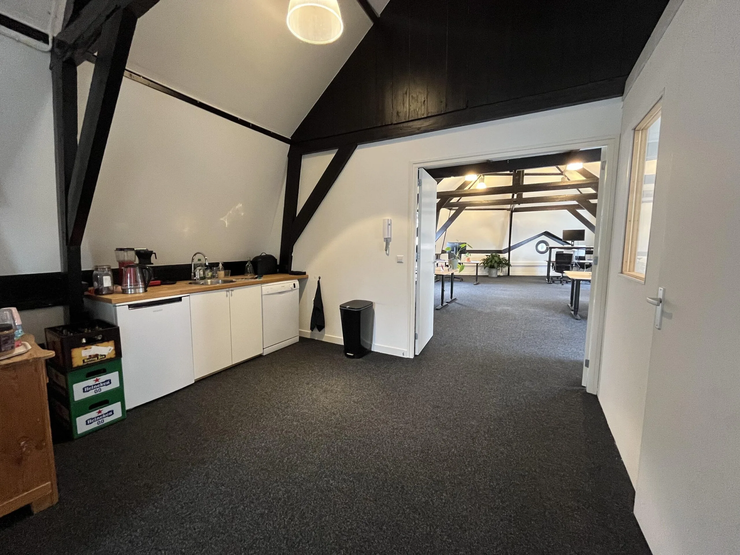 Een kantoorruimte met een kitchenette en open doorgang naar een werkruimte met bureaus en planten.