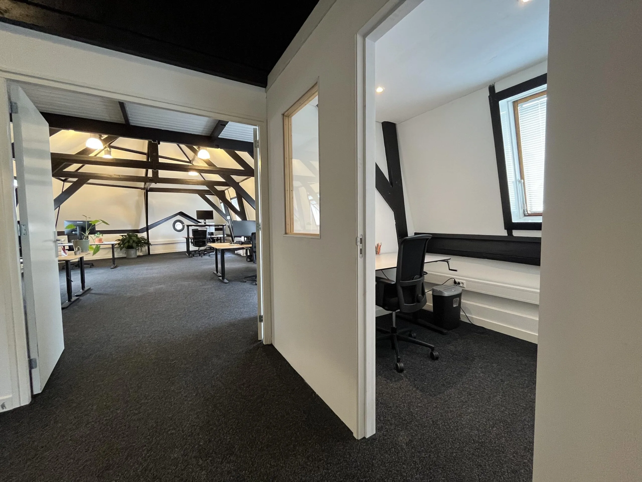 Een moderne, open kantoorruimte met een aparte kamer, donkere balken en bureaus.
