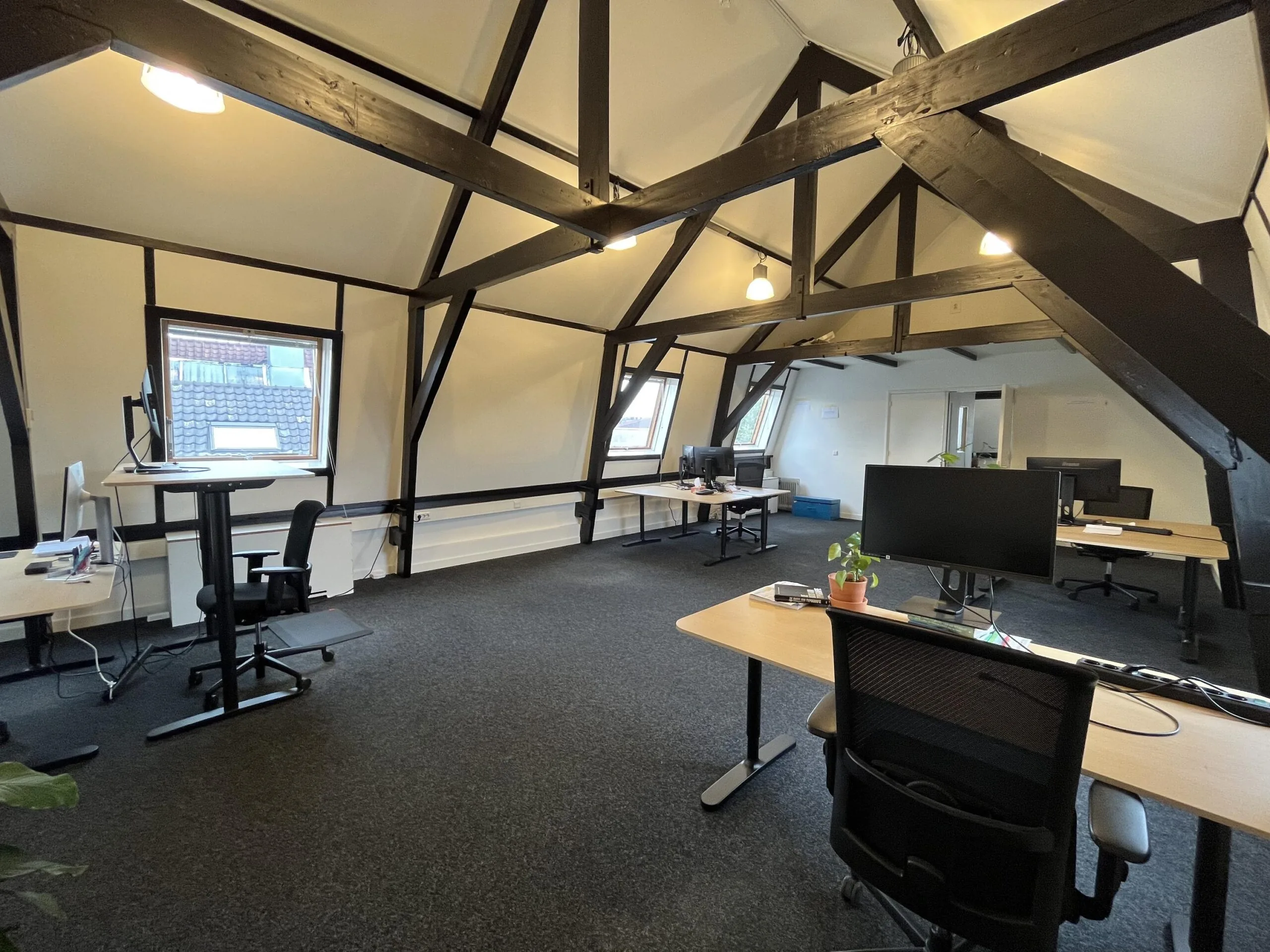 Een kantoorruimte op een zolder met houten balken, bureaus, stoelen en computerwerkplekken.