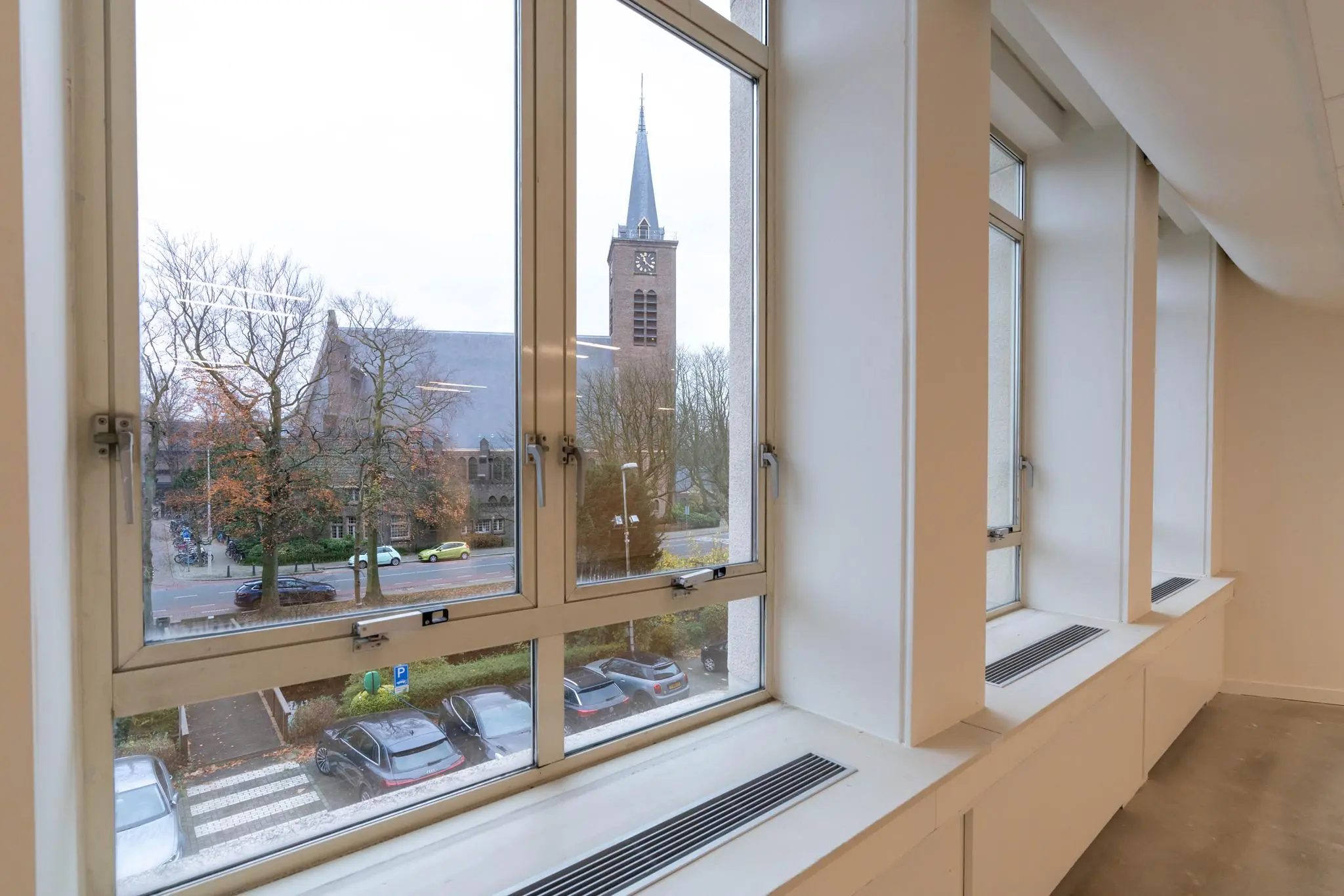 Uitzicht vanuit een modern kantoorraam op de Wassenaarseweg met zicht op een kerk en geparkeerde auto's.