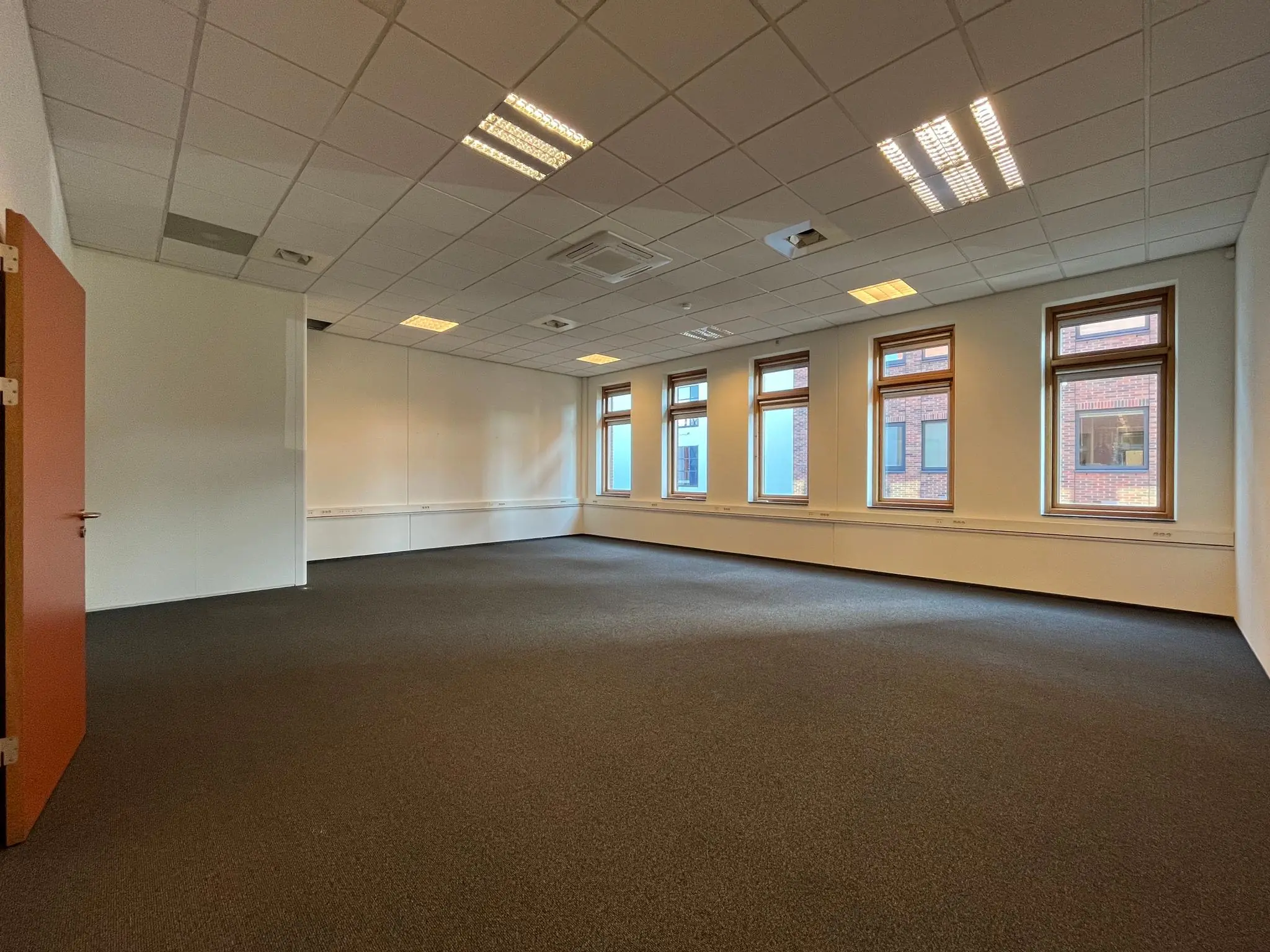 Lege kantoorruimte met systeemplafond, tapijtvloer en meerdere ramen met uitzicht op een bakstenen gebouw.
