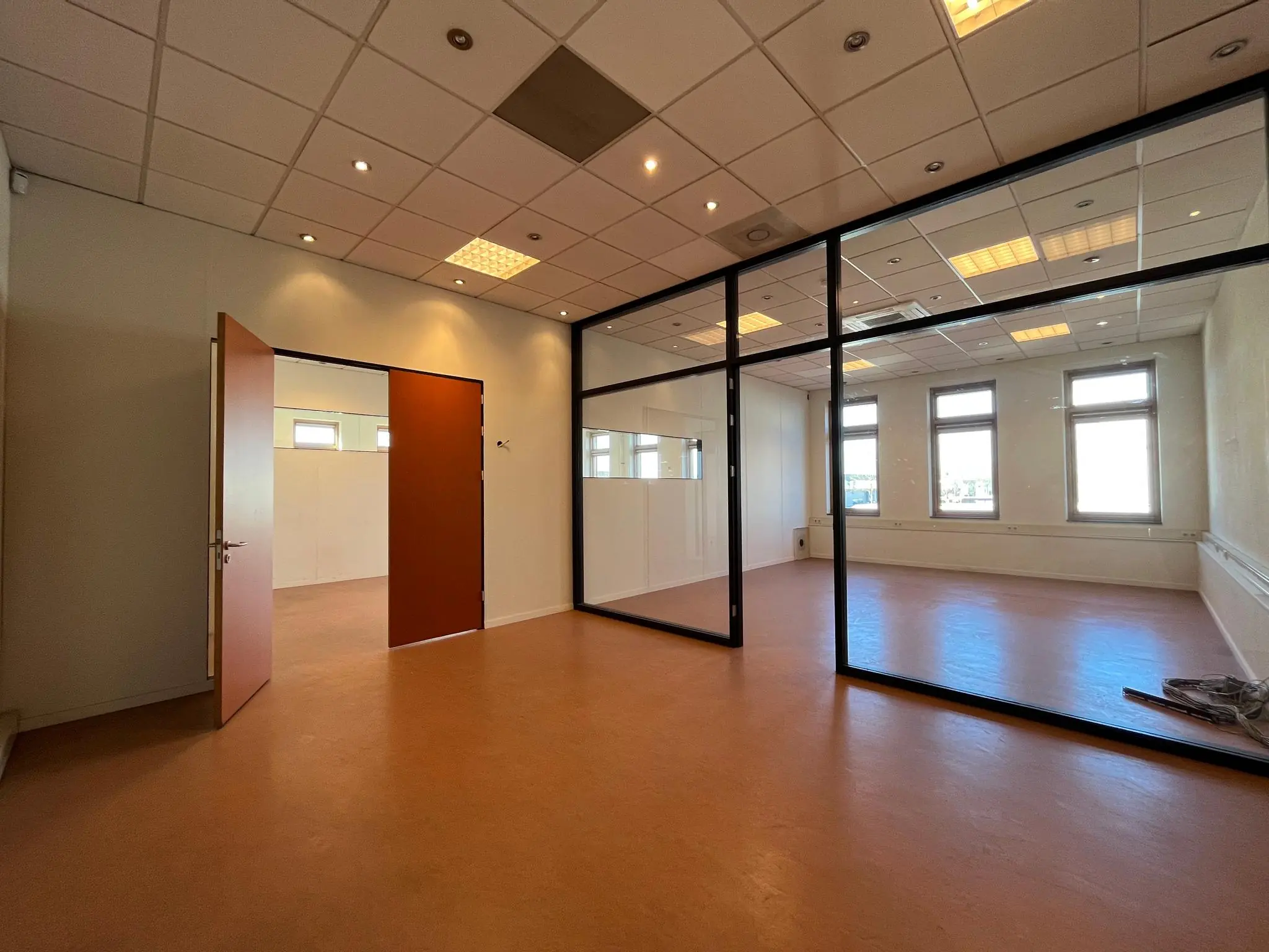 Licht kantoorinterieur met oranjebruine vloer, glazen scheidingswand en openstaande deur.