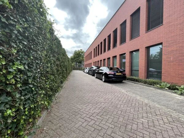 Een rood bakstenen gebouw naast een betegeld pad met auto's en een groene haag op de achtergrond.