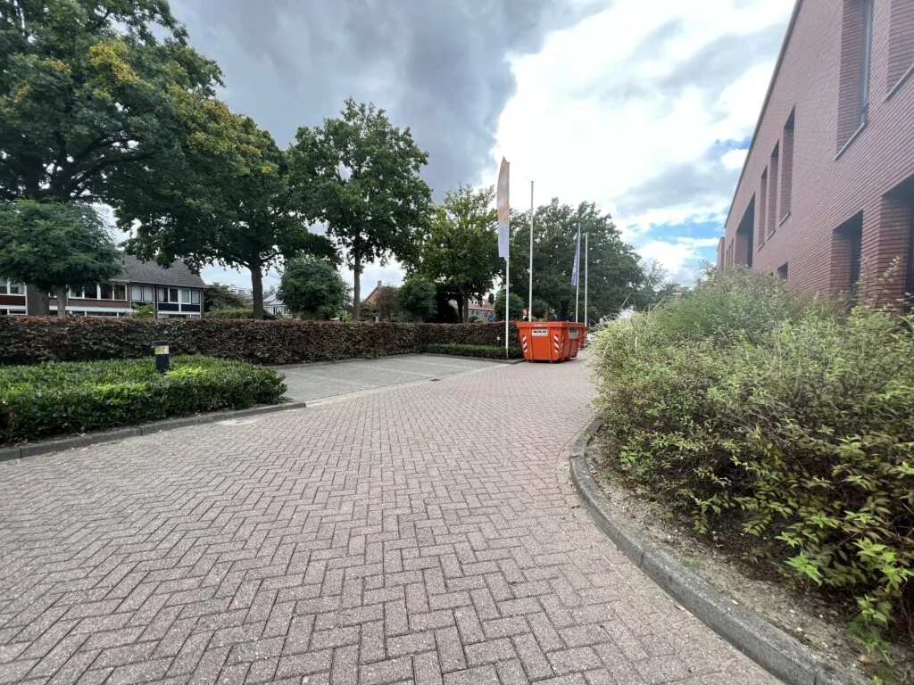 Een rustige straat met een trottoir, omgeven door bomen, een bakstenen gebouw en oranje containers langs de Groenekanseweg.