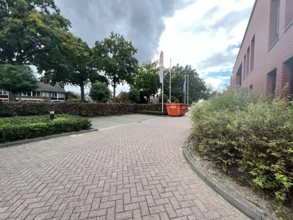 Een rustige straat met een trottoir, omgeven door bomen, een bakstenen gebouw en oranje containers langs de Groenekanseweg.