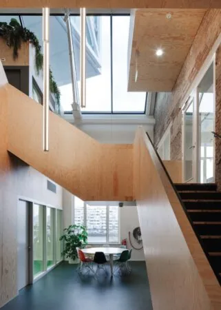 Modern interieur met houten trap, hoge ramen en een vergadertafel met gekleurde stoelen onder een lichtkoepel op de Nassaukade.