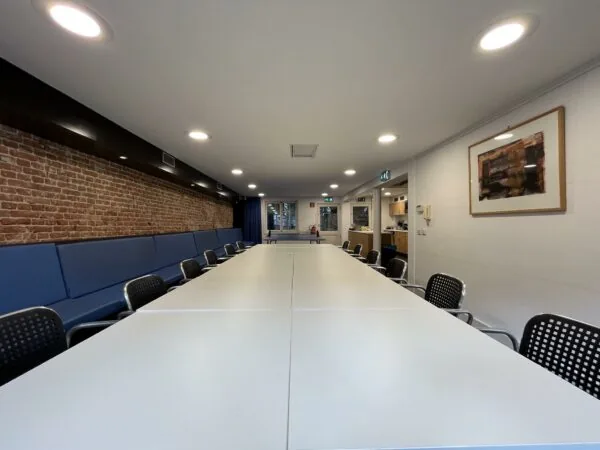 Vergaderruimte aan de Ramstraat met lange witte tafels, zwarte stoelen en een bakstenen muur met blauwe bank.