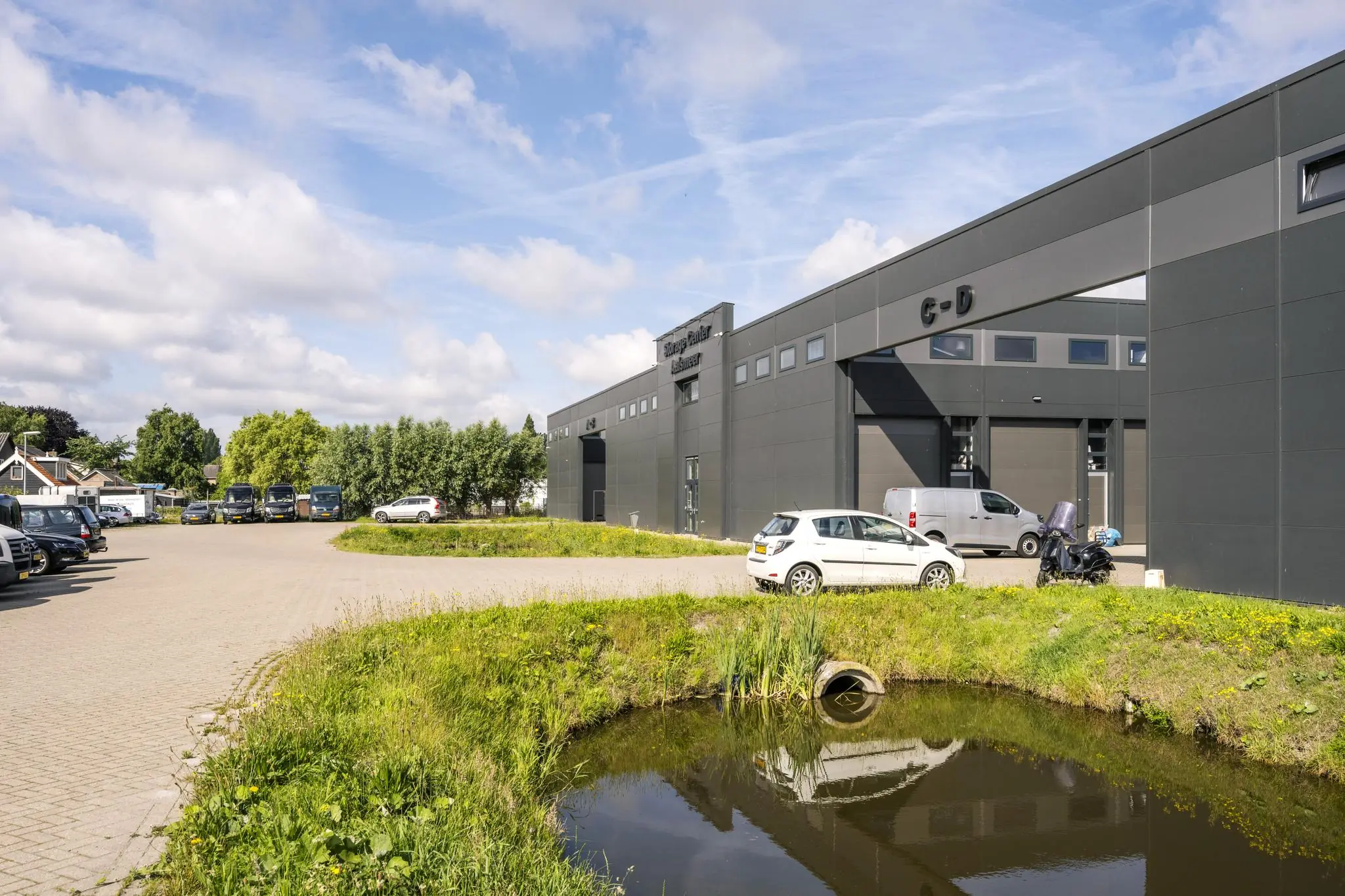 Modern bedrijfsgebouw aan de Aalsmeerderweg met parkeerplaatsen en vijver op de voorgrond.