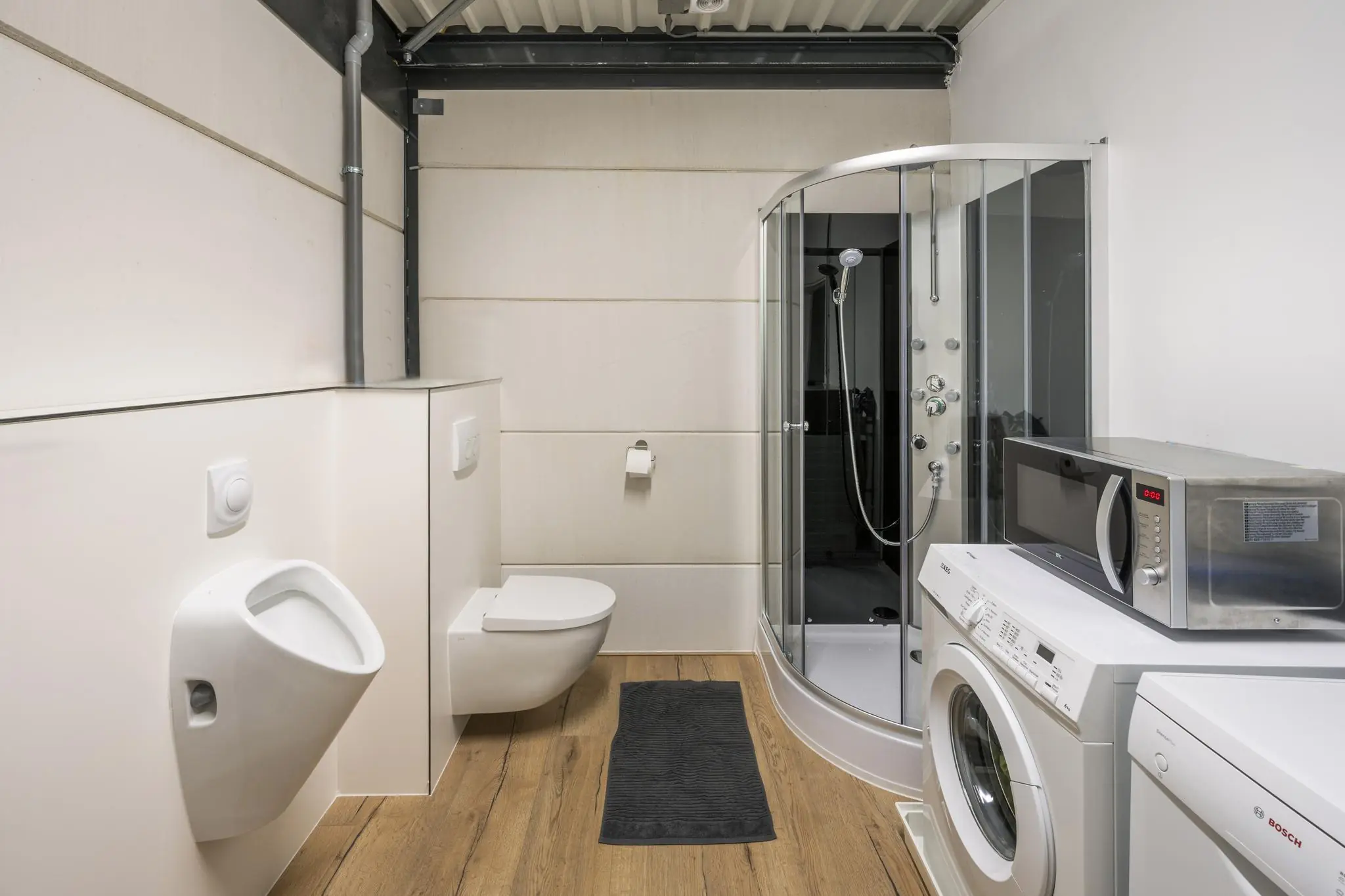 Badkamer met houten vloer, voorzien van urinoir, toilet, douchecabine, wasmachine, droger en magnetron aan de Aalsmeerderweg.