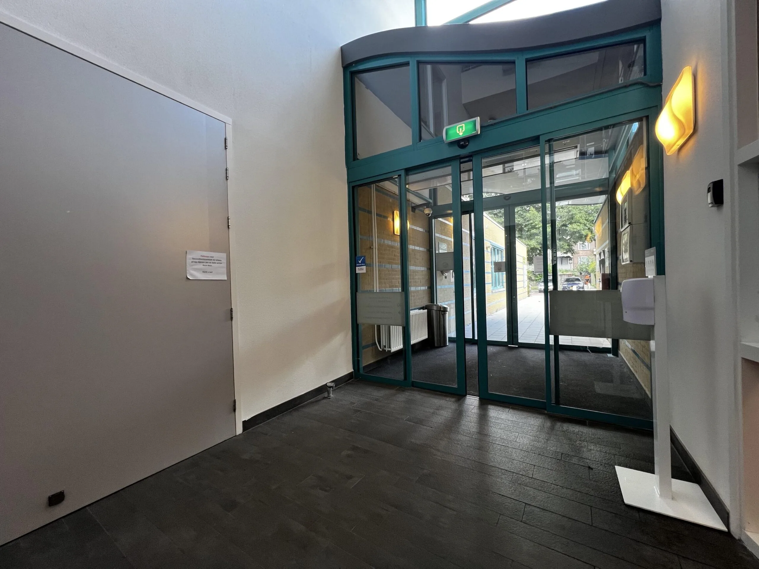 Ingang van een modern gebouw met glazen schuifdeuren en een handdesinfectiestation.